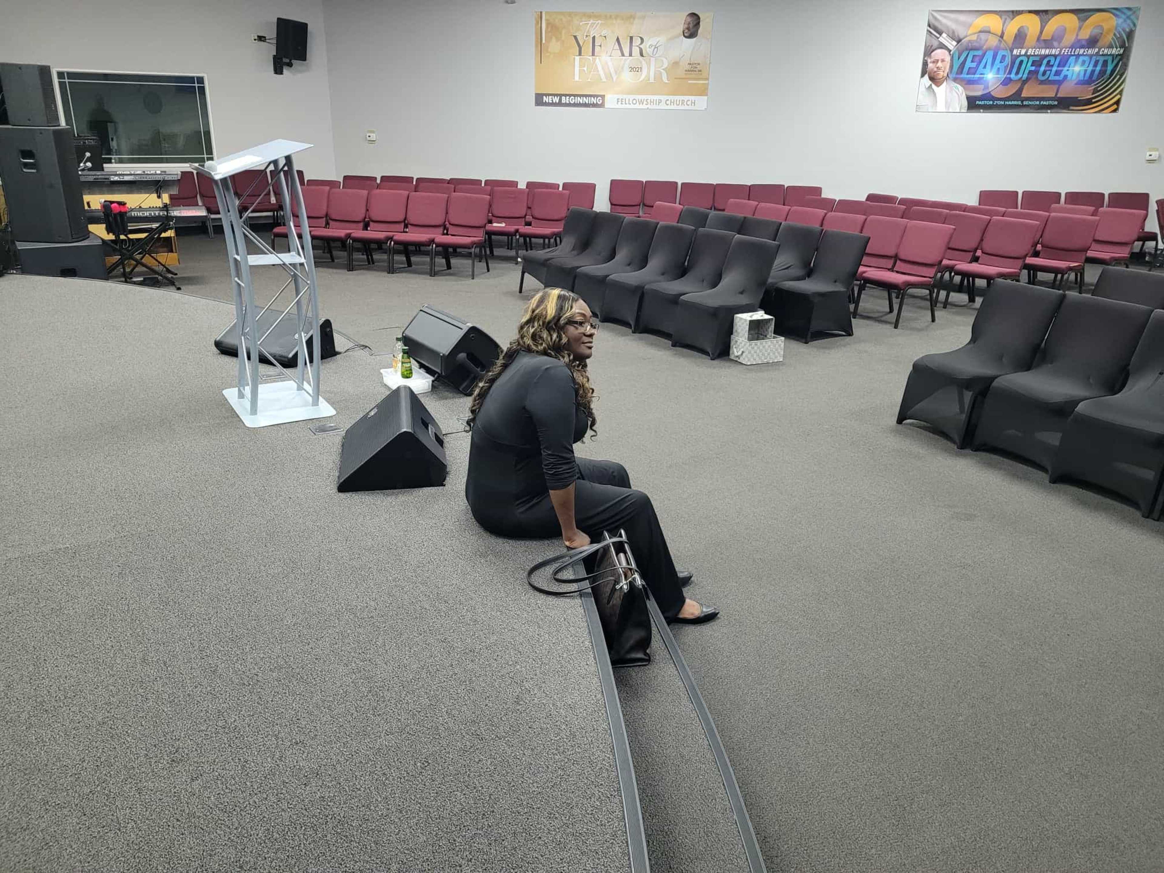 A woman sits on the floor of a nearly empty church auditorium with rows of chairs in the background.