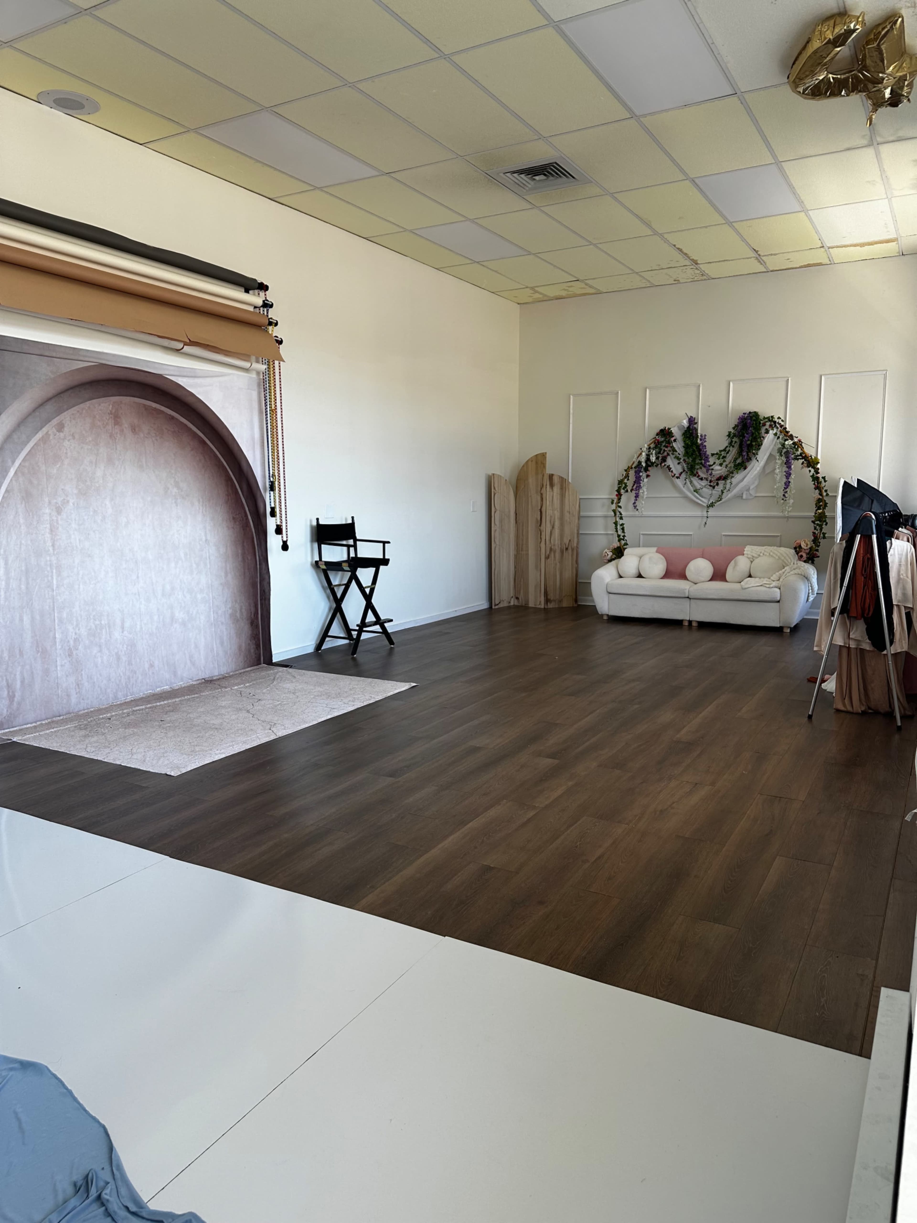 The image shows a spacious photography studio with a wooden floor, a backdrop, a chair, a sofa adorned with decorative pillows, and some greenery.