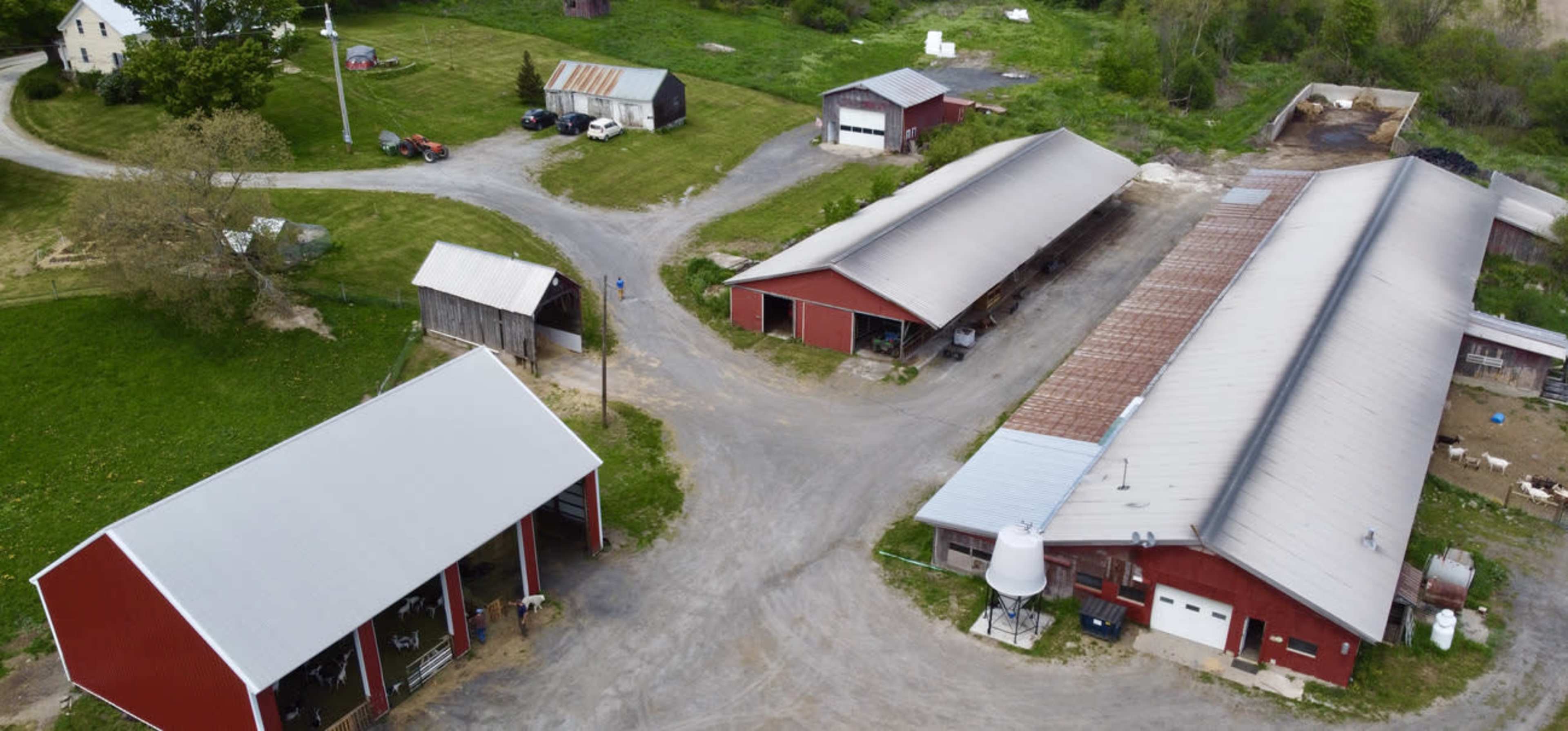 An aerial view of a rural farm with multiple red barns, a circular driveway, and several vehicles parked nearby.