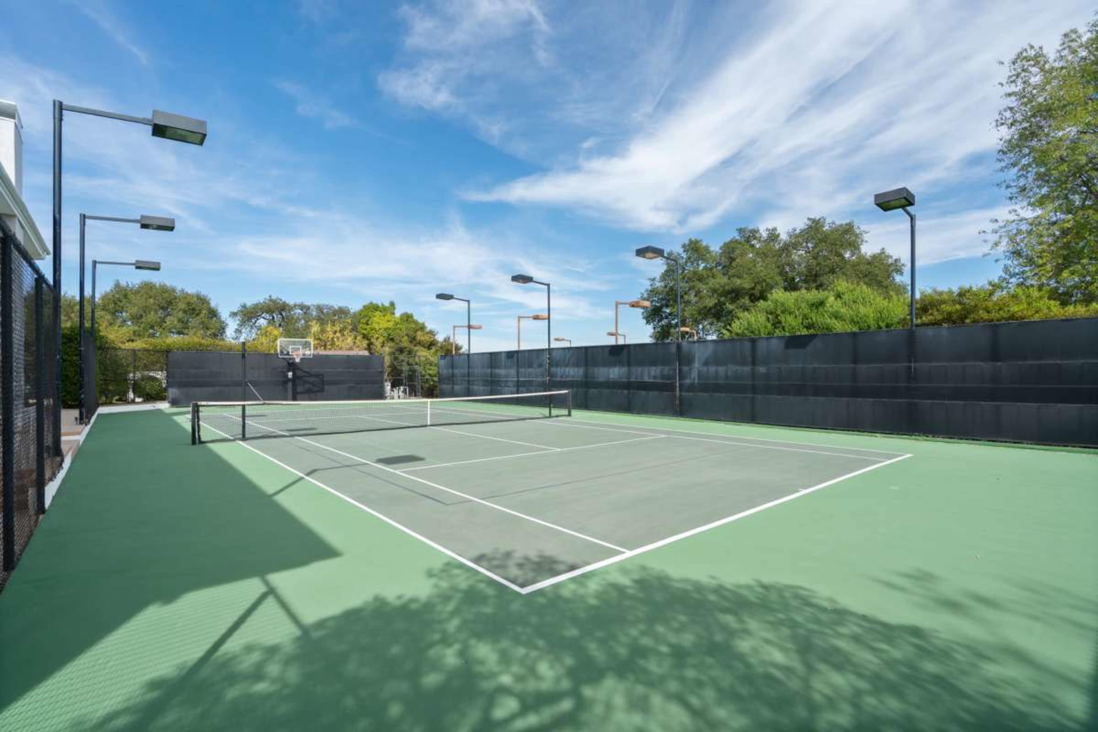 The image shows an empty outdoor tennis court surrounded by black fencing and trees under a blue sky.