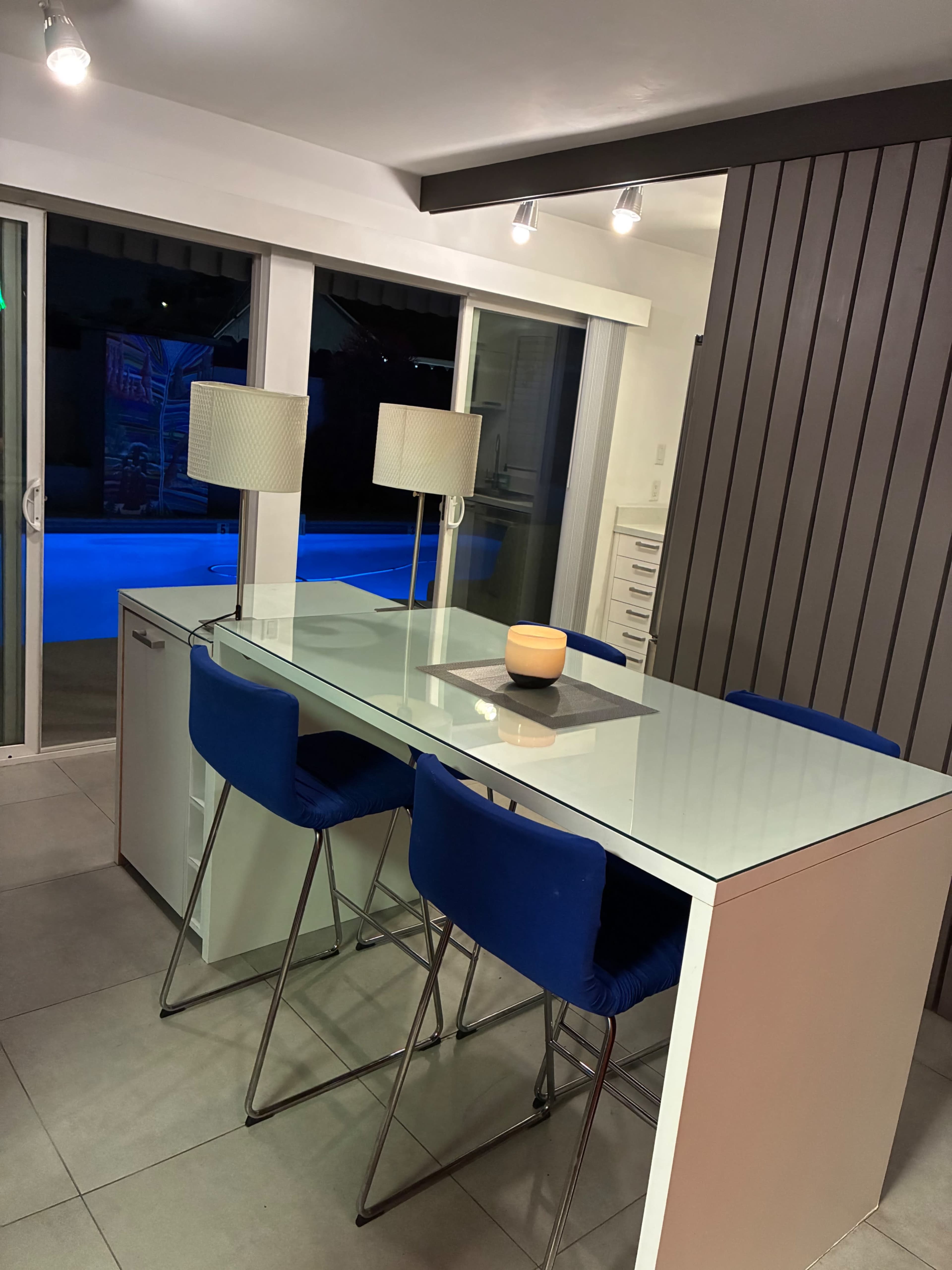 The image shows a modern dining area with a glass-top table, blue chairs, and lamps, overlooking a pool through large windows.