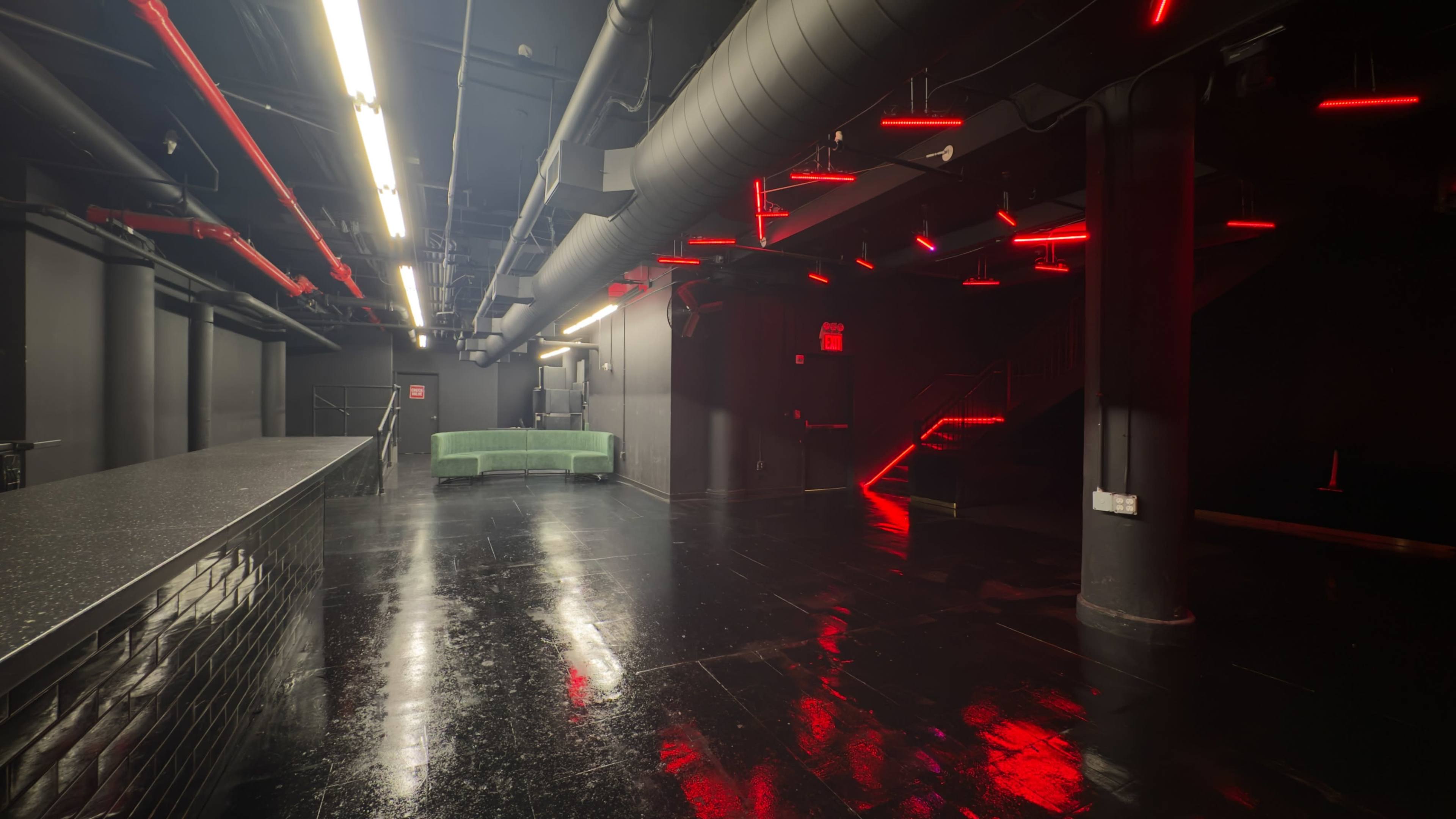 The interior of a dimly lit venue features black floors, exposed ventilation, and red neon lighting accenting the walls and stairway.