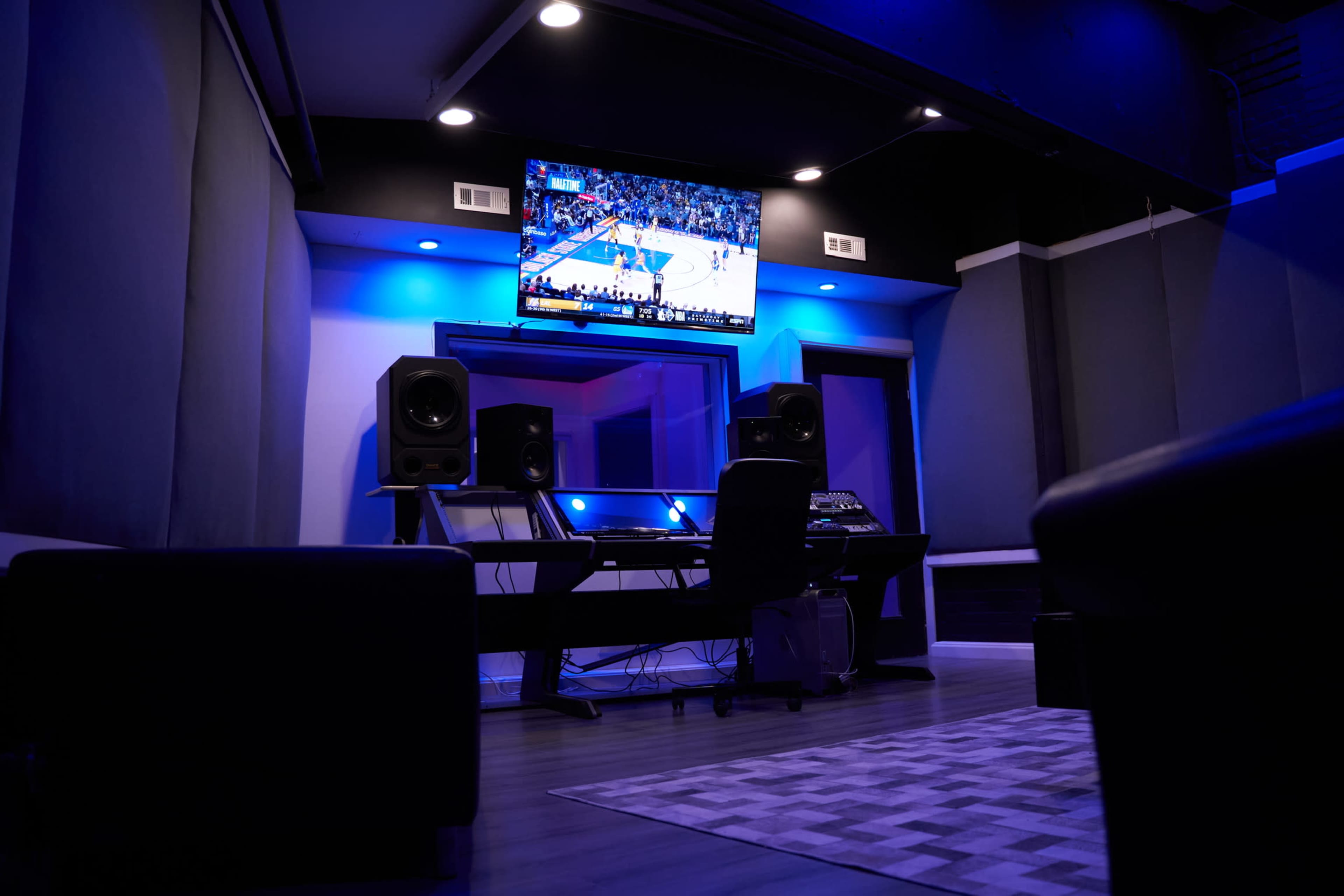 A modern recording studio features a large screen displaying a basketball game, with audio equipment and blue lighting.