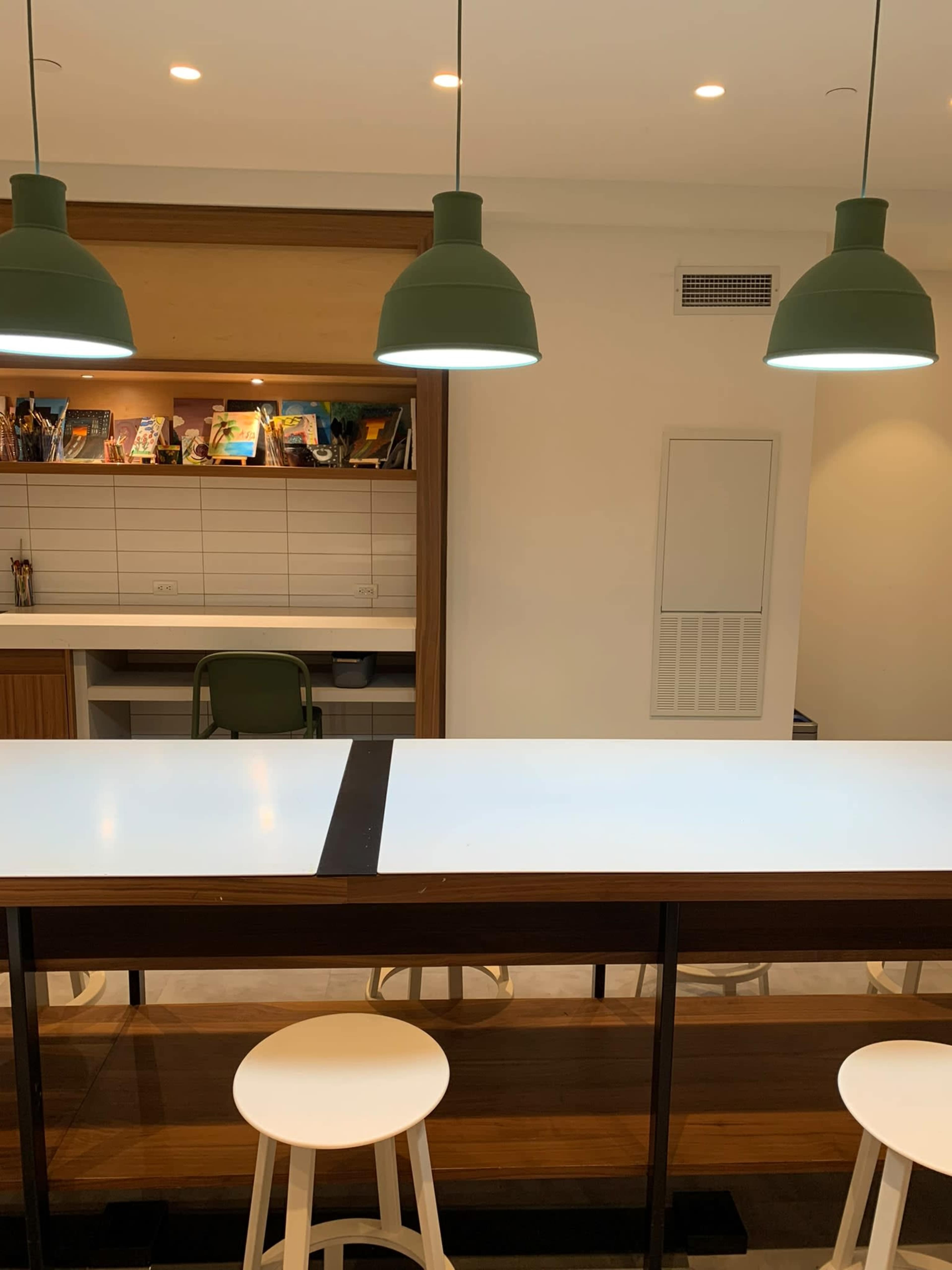 A modern kitchen area features a long, white countertop with bar stools and green pendant lights above.