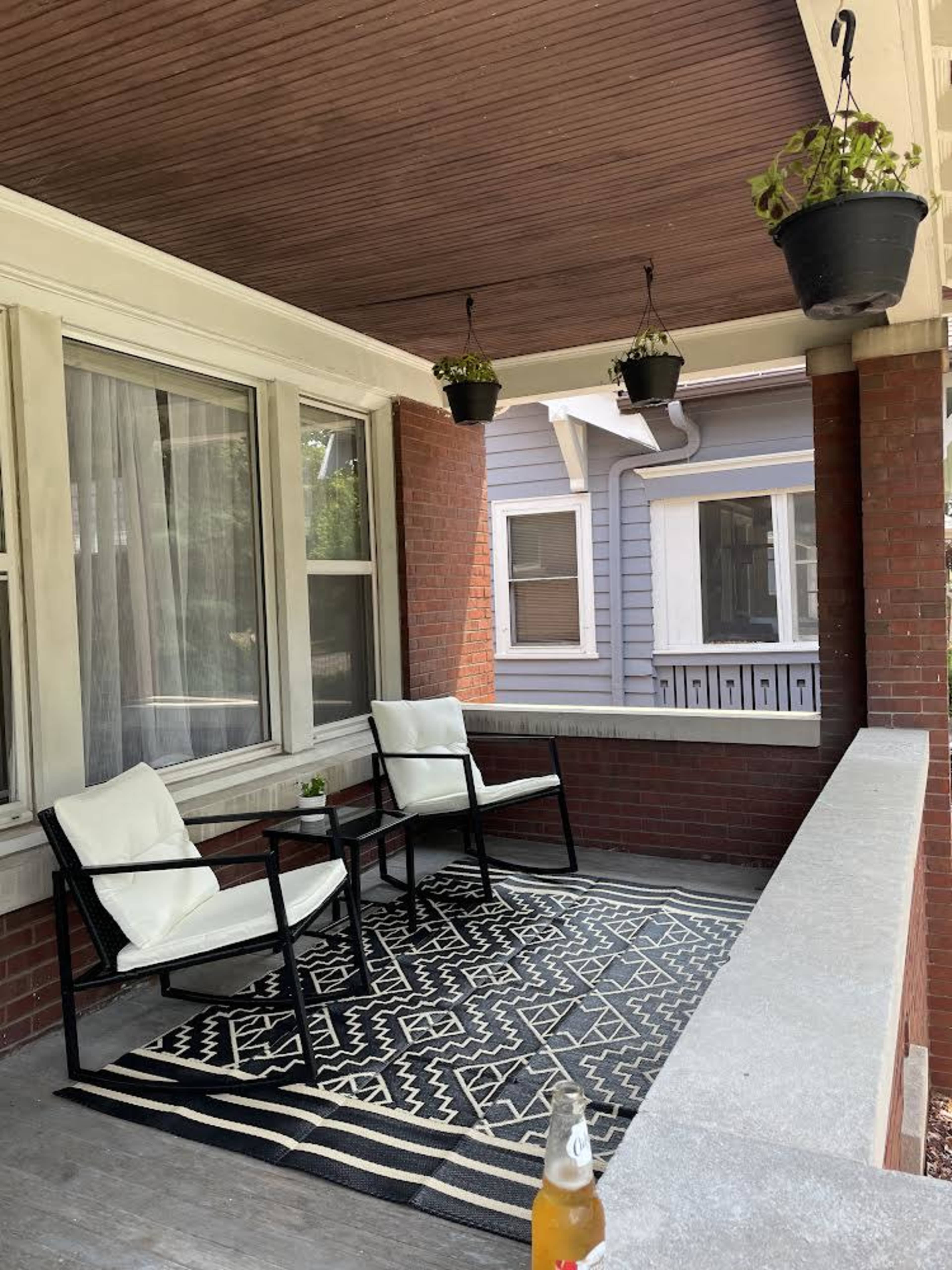 A covered porch with two white chairs and a small table on a patterned rug, surrounded by brick walls and adorned with hanging potted plants.