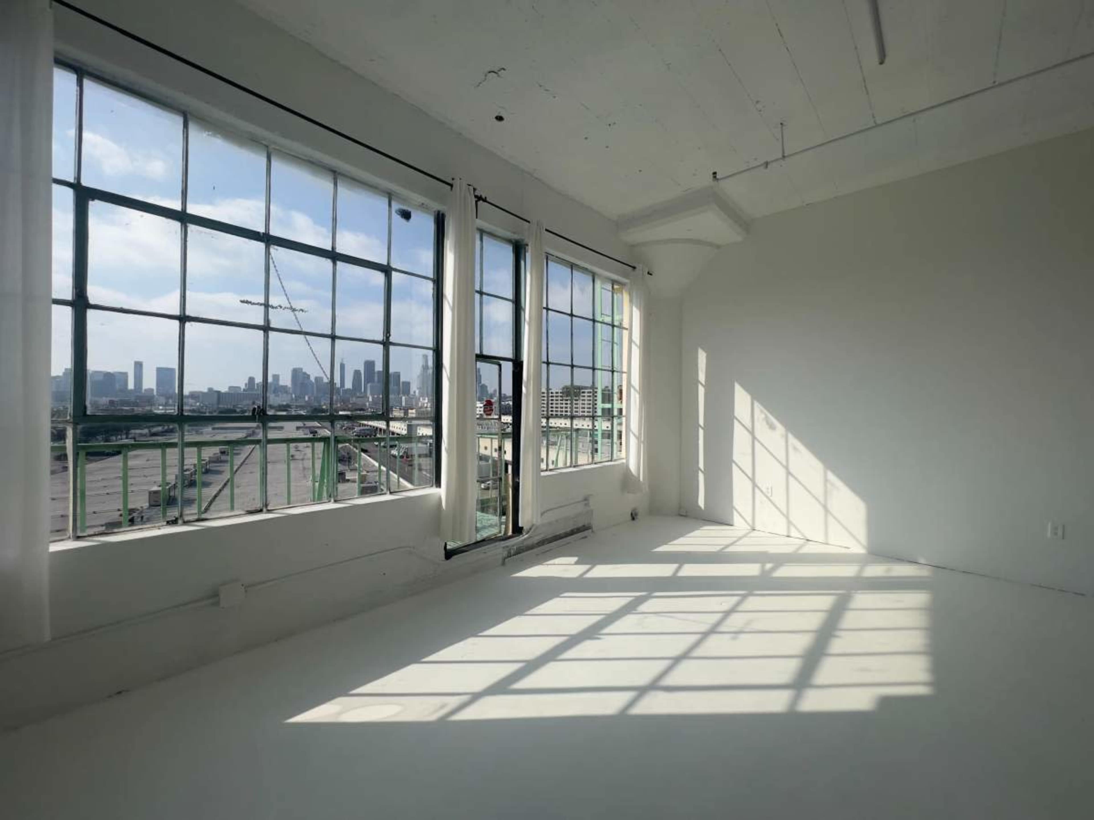 A sunlit, empty room with large windows overlooks a city skyline.