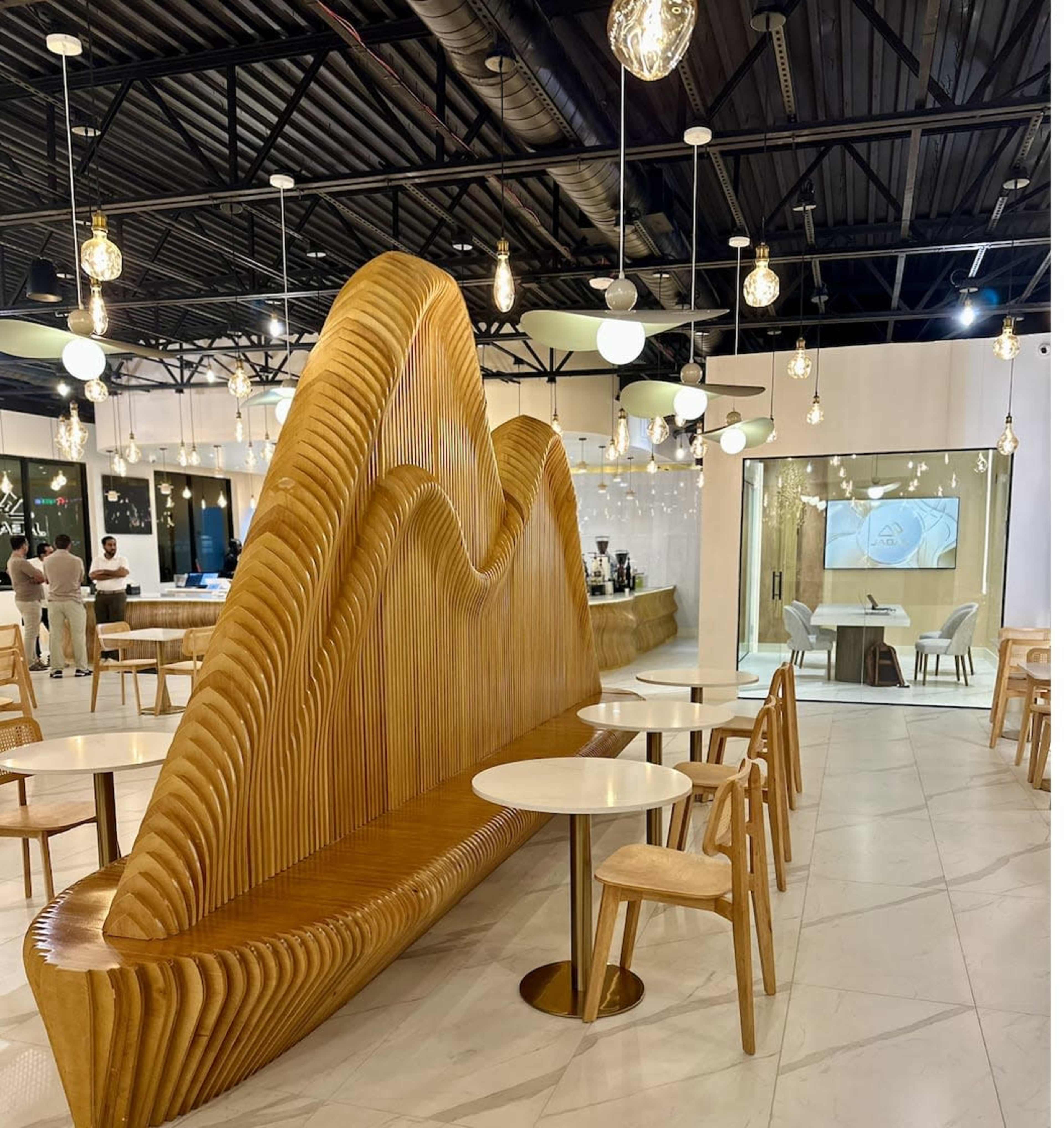 The image shows a modern café interior with a distinctive wooden wave-shaped bench, white tables, and pendant lighting.