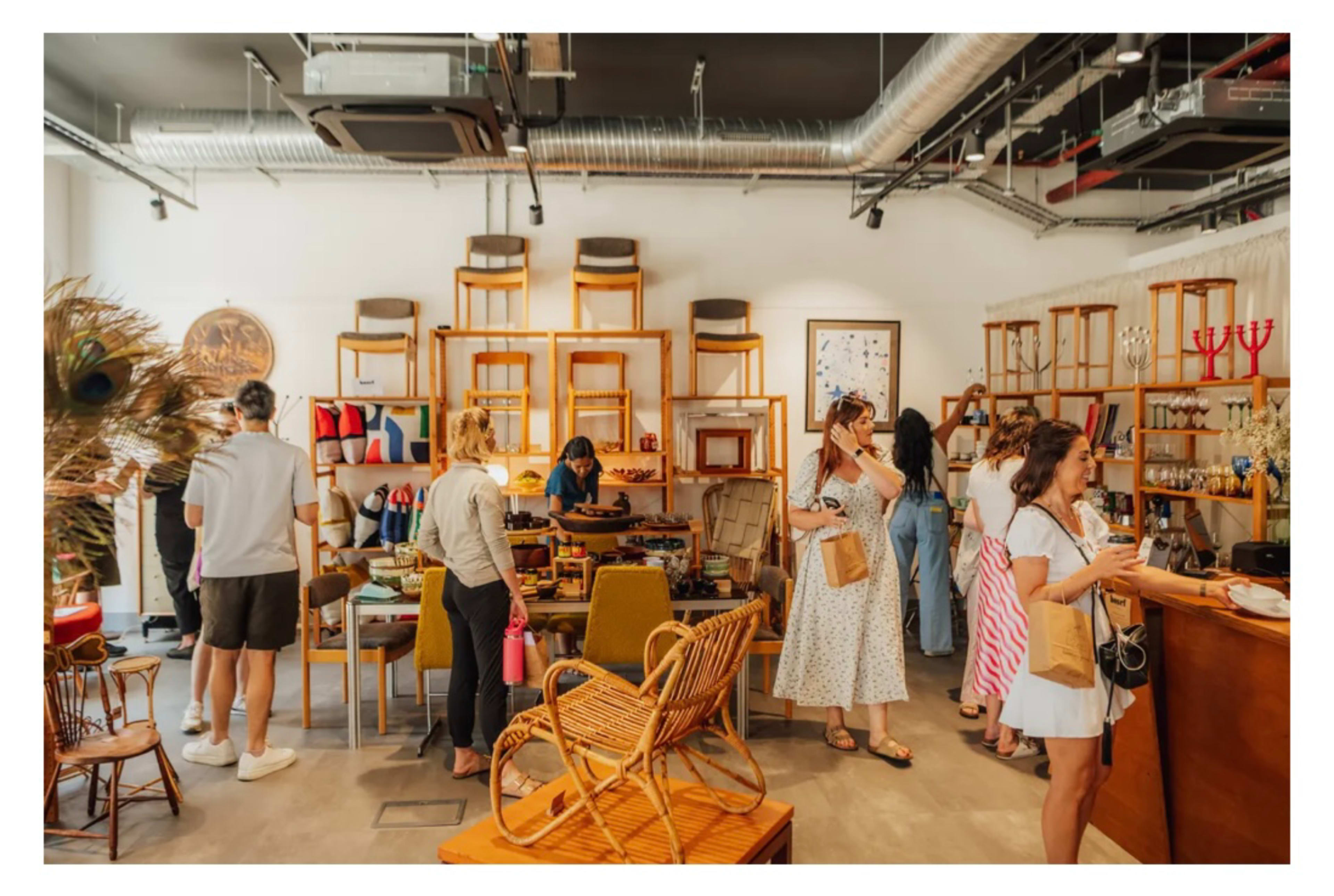 A busy retail space features wooden furniture and various artisanal products, with several customers browsing and interacting inside the store.