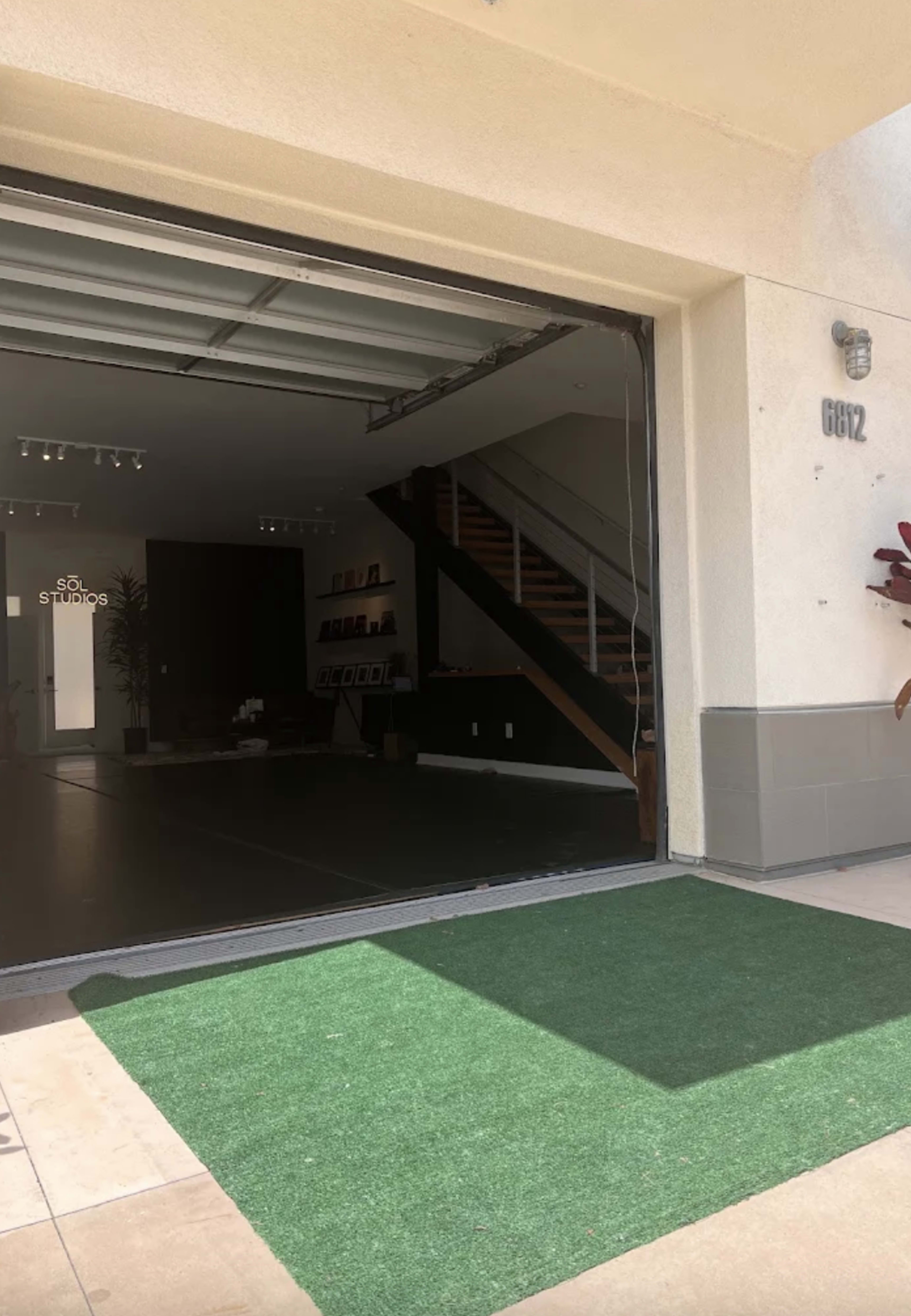 The image shows the entrance of a modern studio with a large open garage door, revealing a staircase and minimalist interior design.