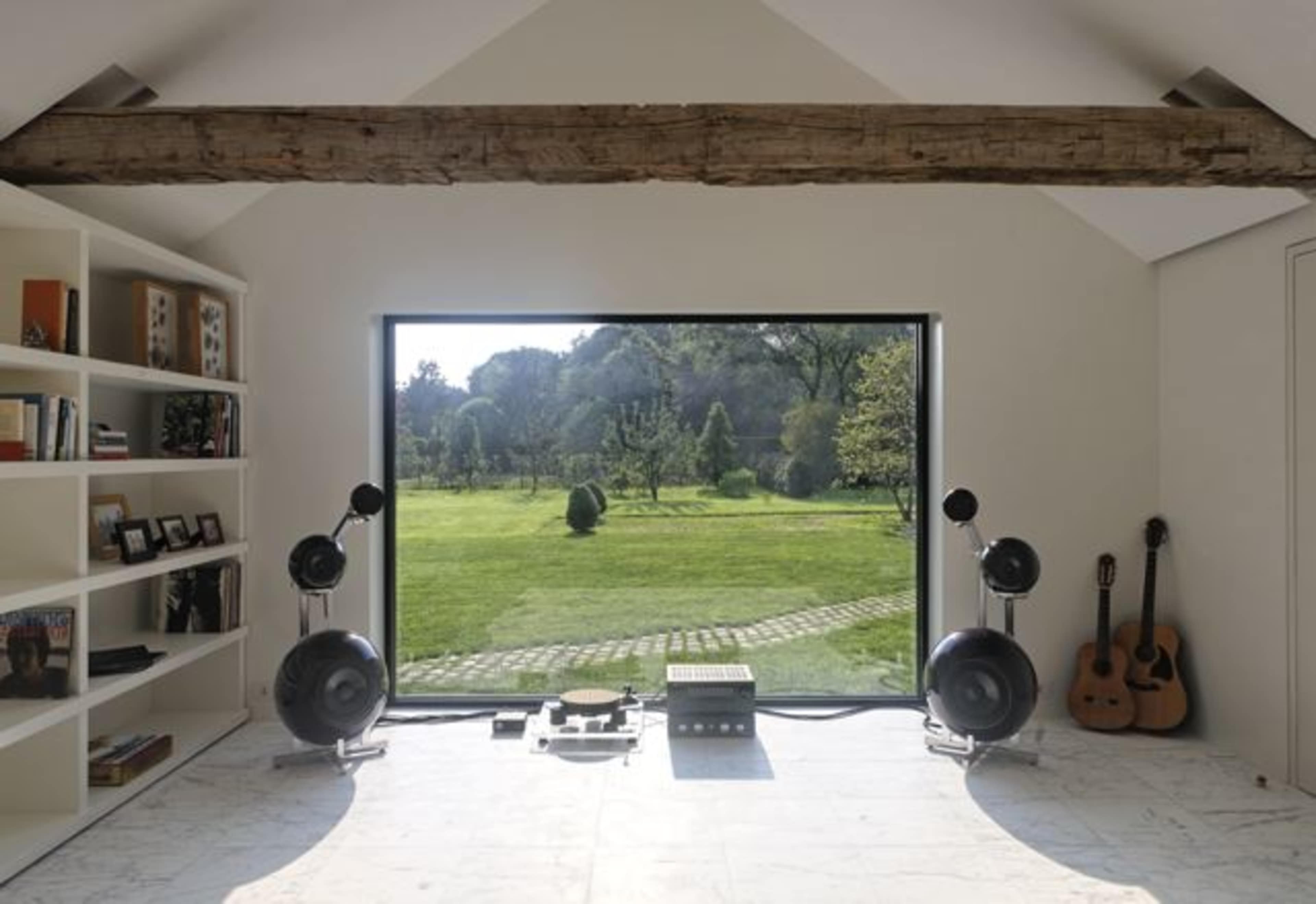 The image shows a minimalist room with a large window overlooking a garden, featuring shelves filled with books and photographs, and a music setup with speakers, a turntable, and a guitar.