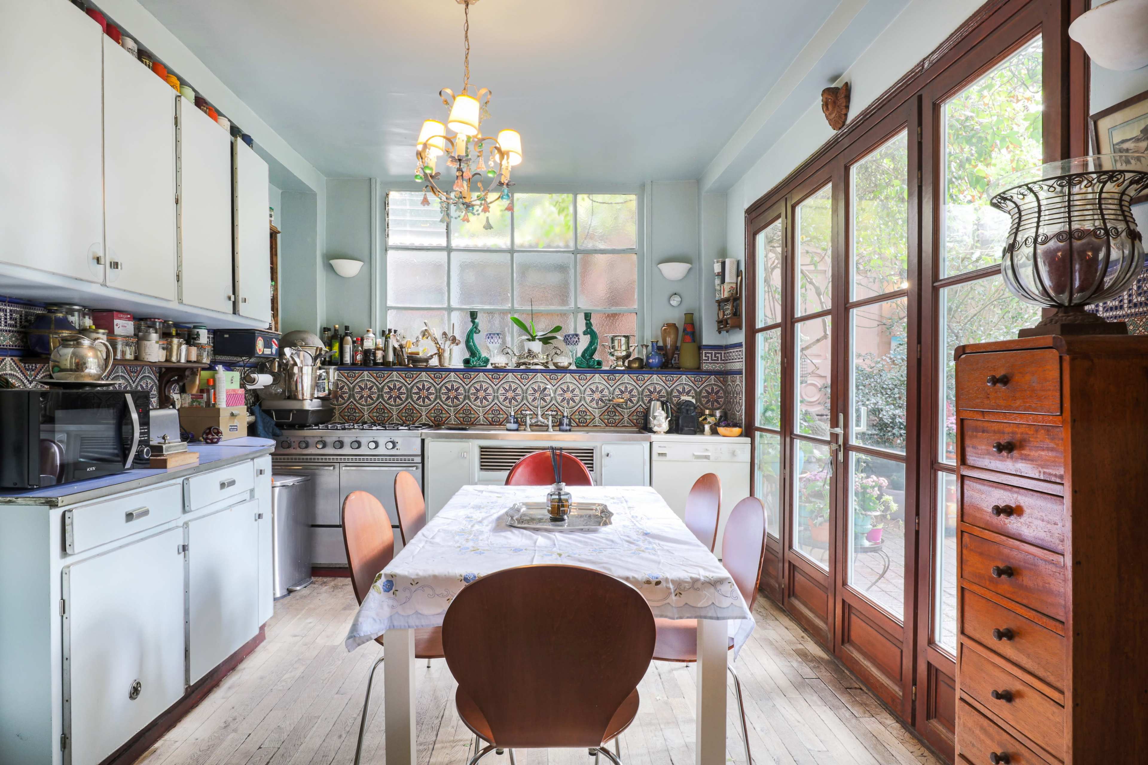 A kitchen features a central table surrounded by chairs, with cabinets and appliances along the walls and large glass doors leading to a garden.