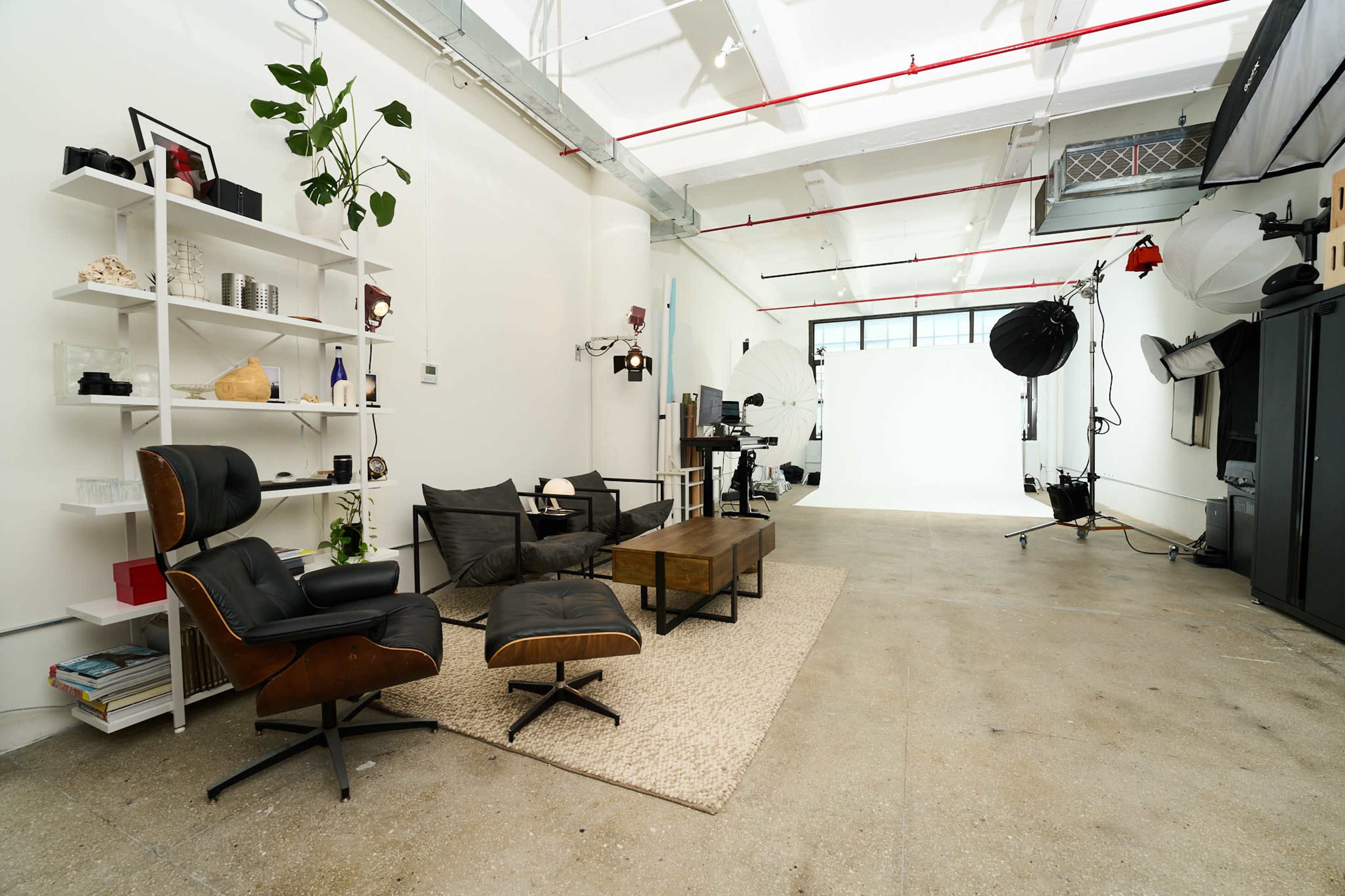The image shows a spacious studio with modern furniture, including a black chair and a wooden coffee table, set against a backdrop of photography equipment.