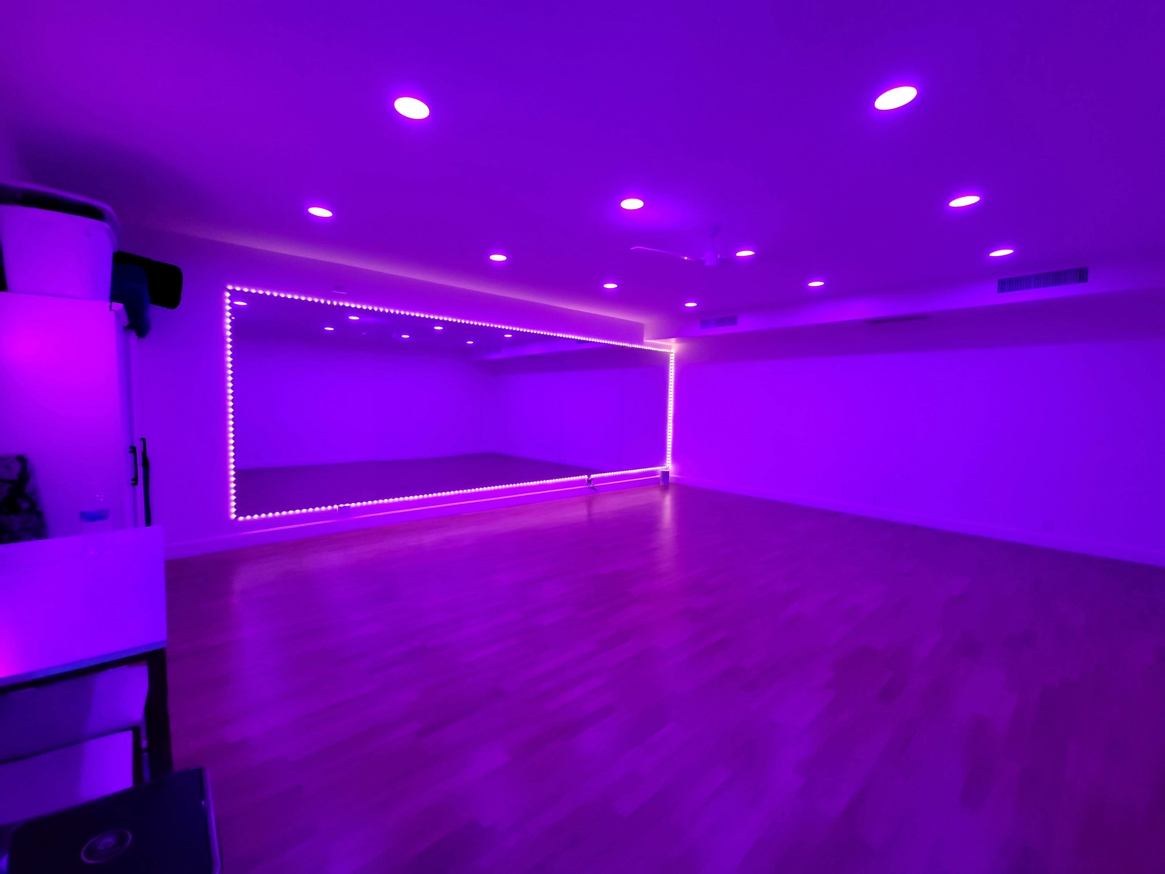 A spacious room with wooden floors, illuminated by purple LED lights and featuring a large mirror on one wall.