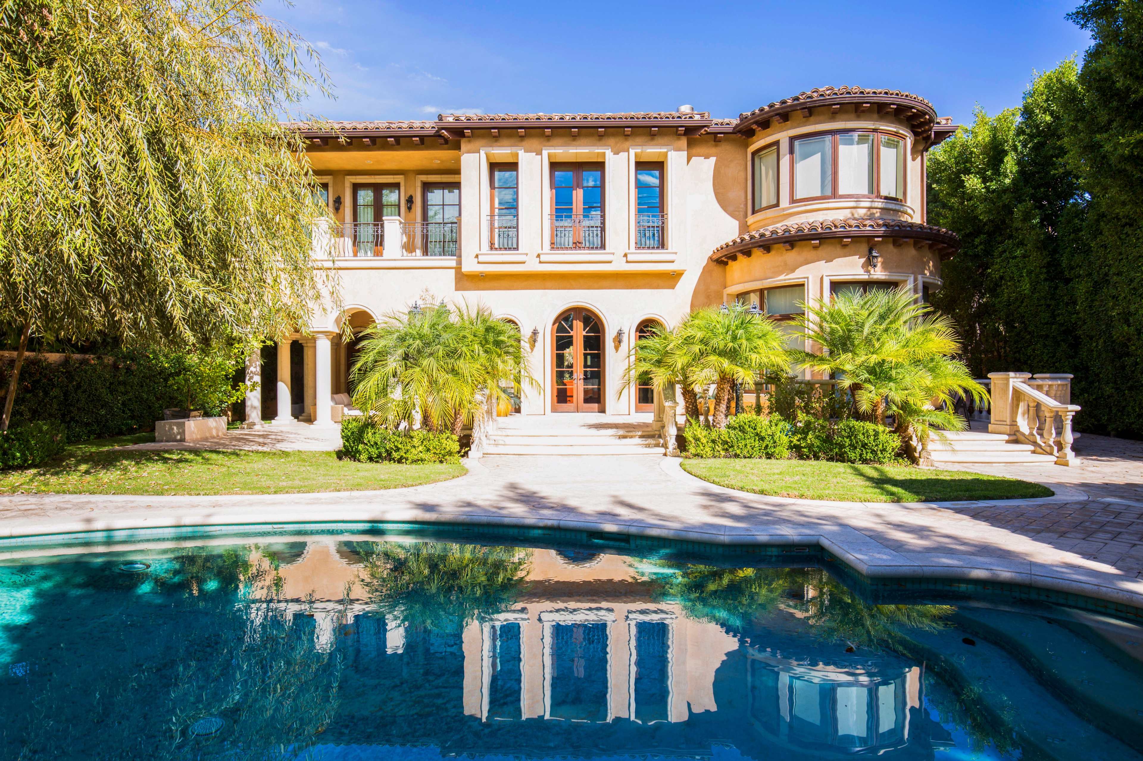 A large, two-story house with arched windows and a swimming pool in the foreground, surrounded by palm trees and well-maintained landscaping.