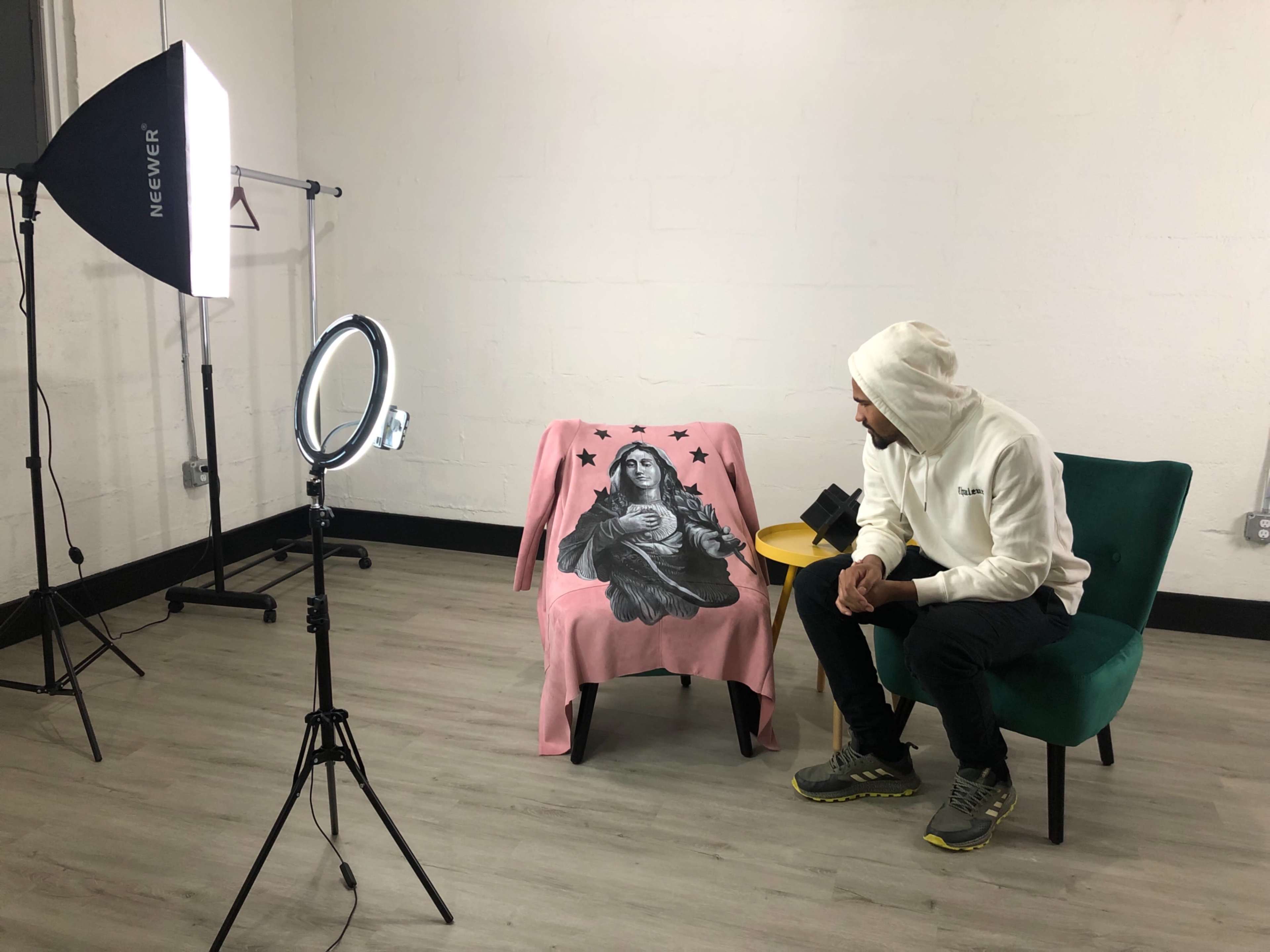A man in a white hoodie sits beside a green chair draped with a pink blanket featuring a design of a figure, with a ring light and a tripod in the background.