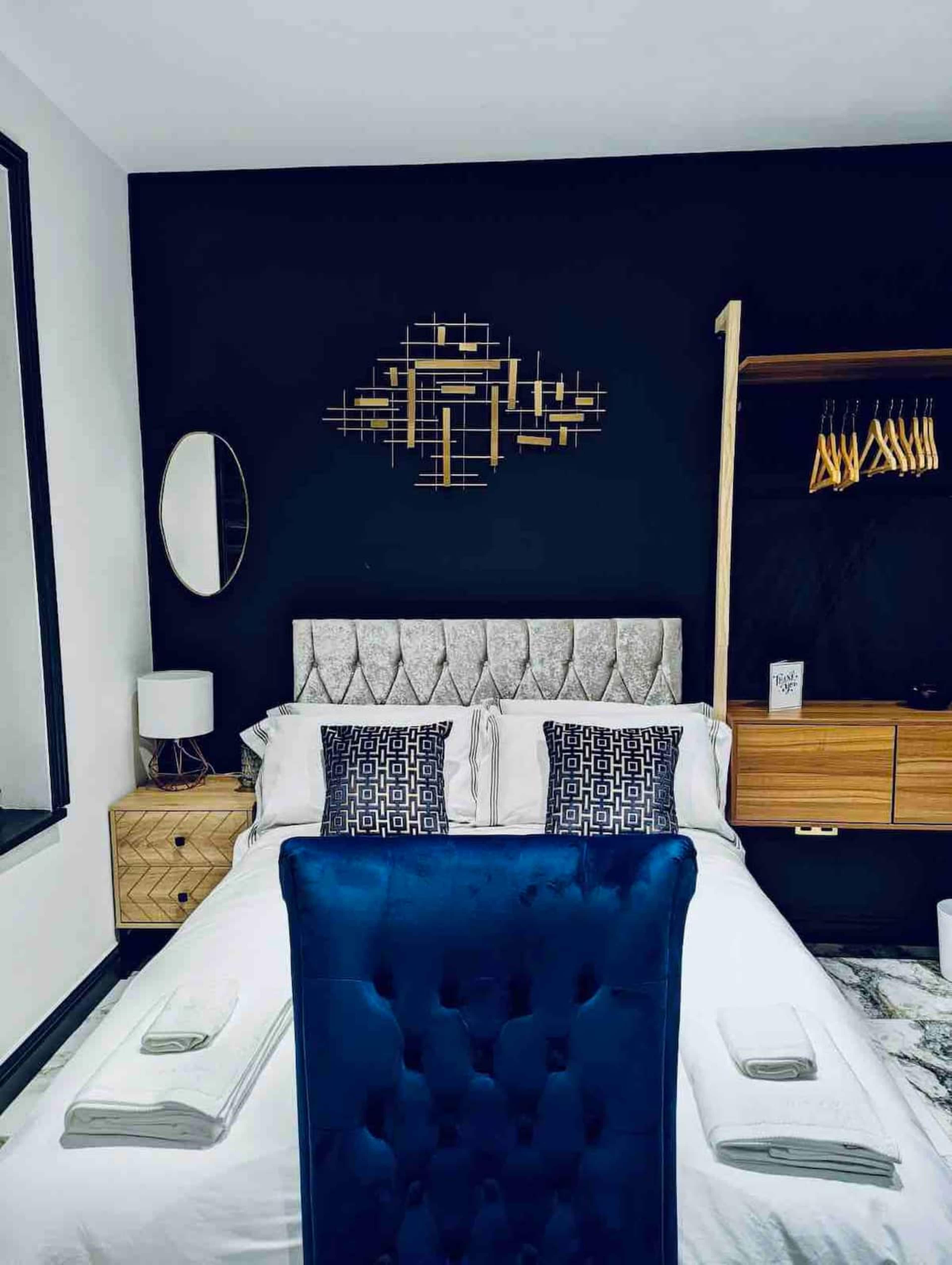 The image shows a modern bedroom featuring a bed with a tufted headboard, a decorative wall clock, and a wooden wardrobe with hangers.