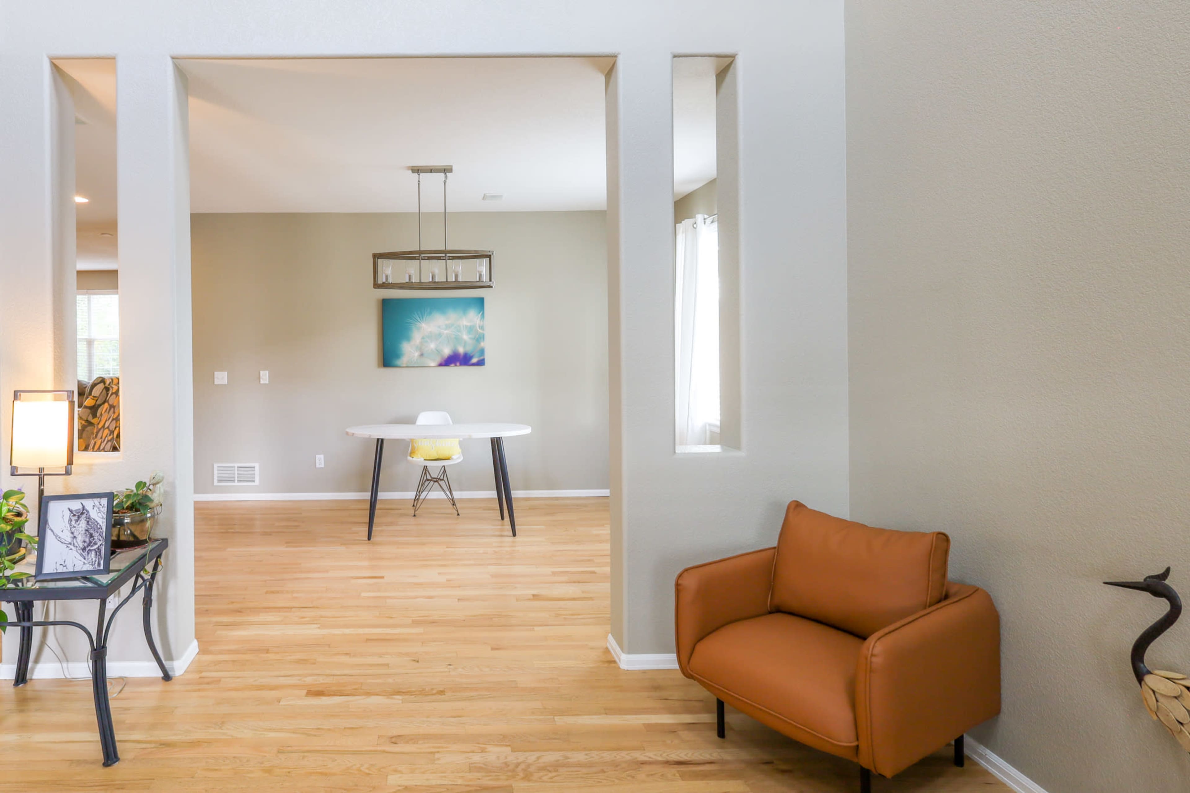 A living space with a small brown chair in the foreground and a dining area featuring a round white table and modern yellow chair in the background, framed by a light-colored wall.