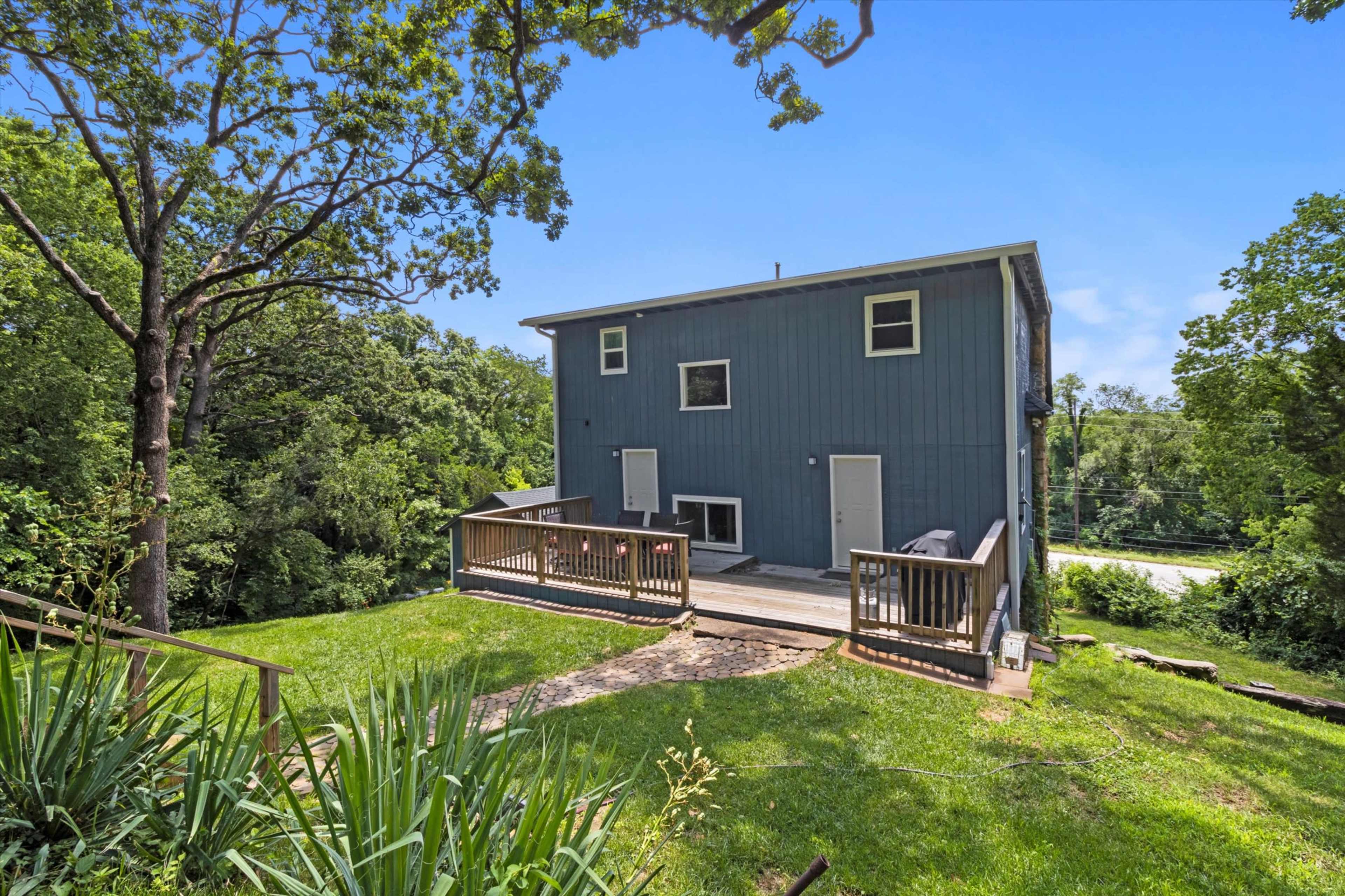 A two-story blue house is situated on a grassy hill with a deck overlooking a wooded area.