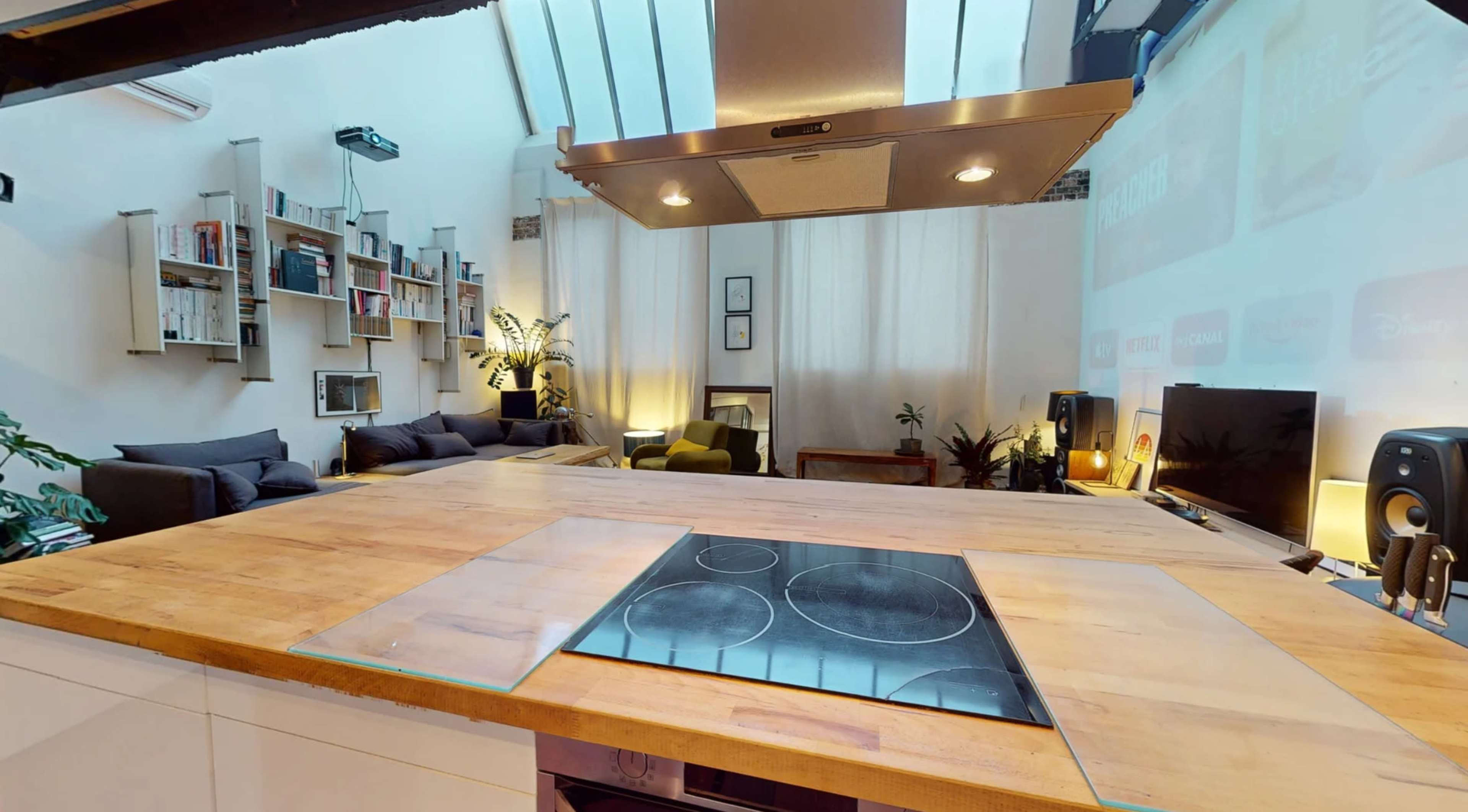 A well-lit kitchen features a large wooden island with an integrated cooktop, surrounded by contemporary living room furniture and plants.