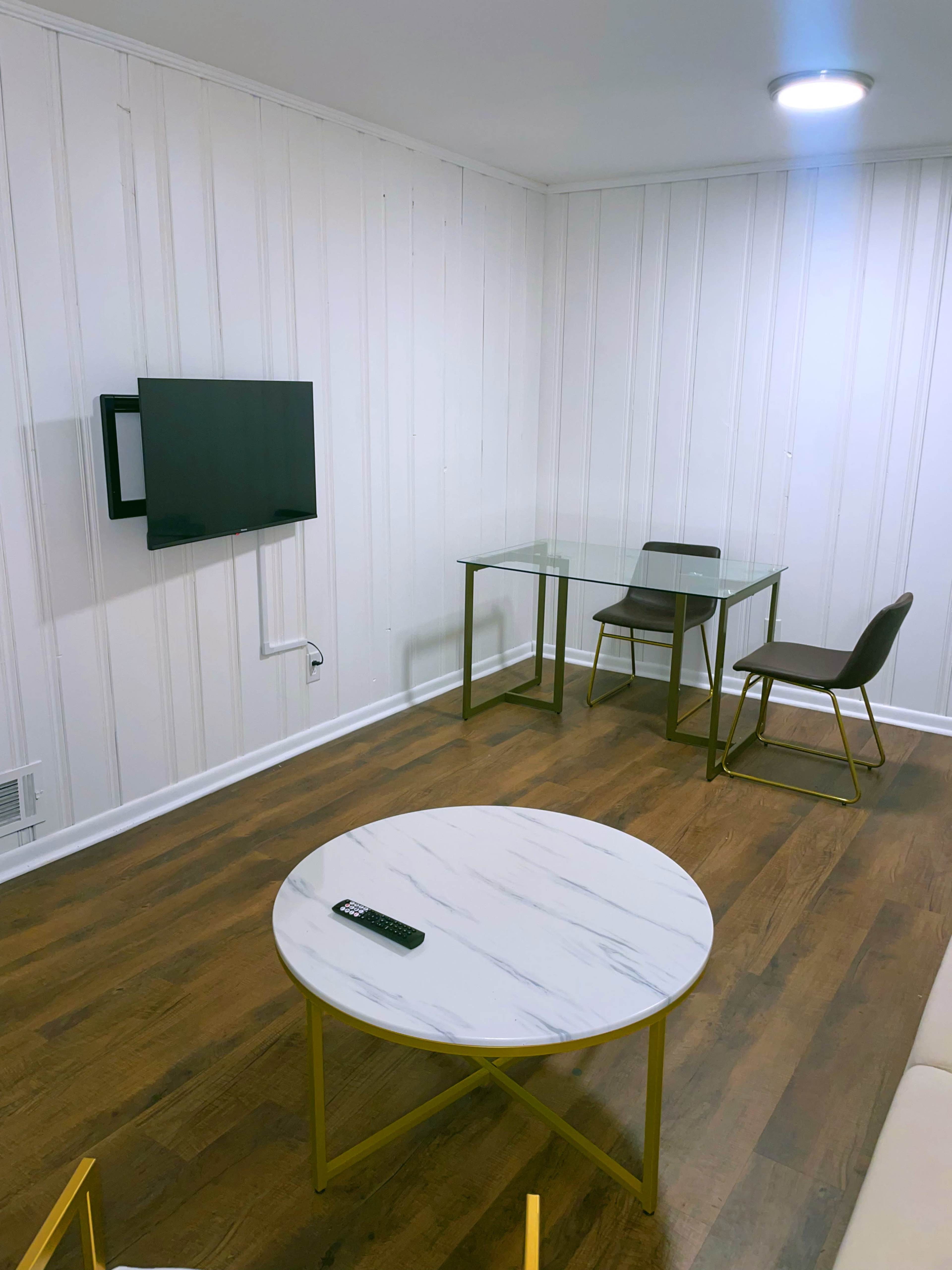 A small room with white paneled walls featuring a television mounted on the wall, a glass table with chairs, and a round marble coffee table.