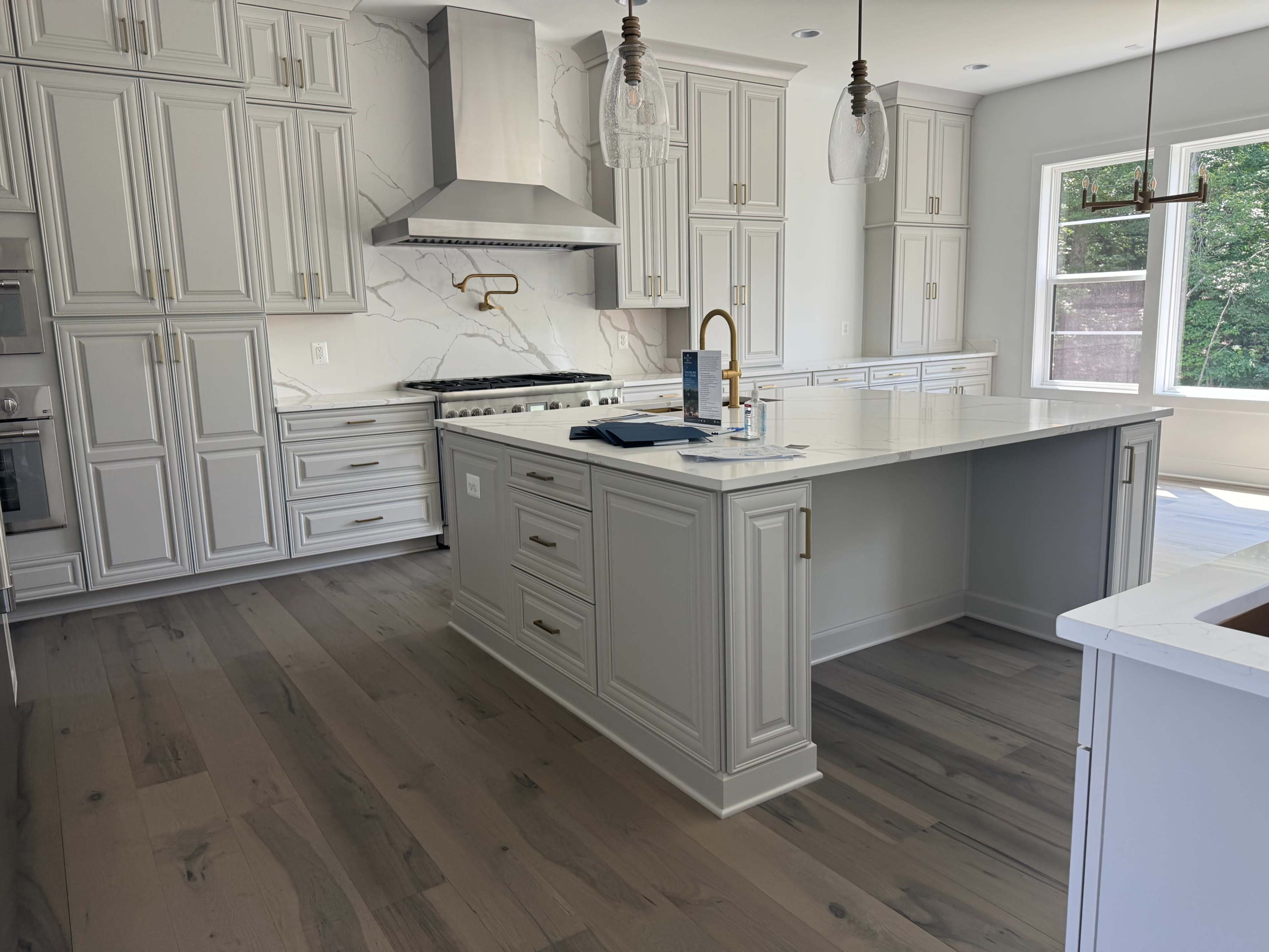 The image shows a modern kitchen with light gray cabinetry, a large island, and a stainless steel range hood.