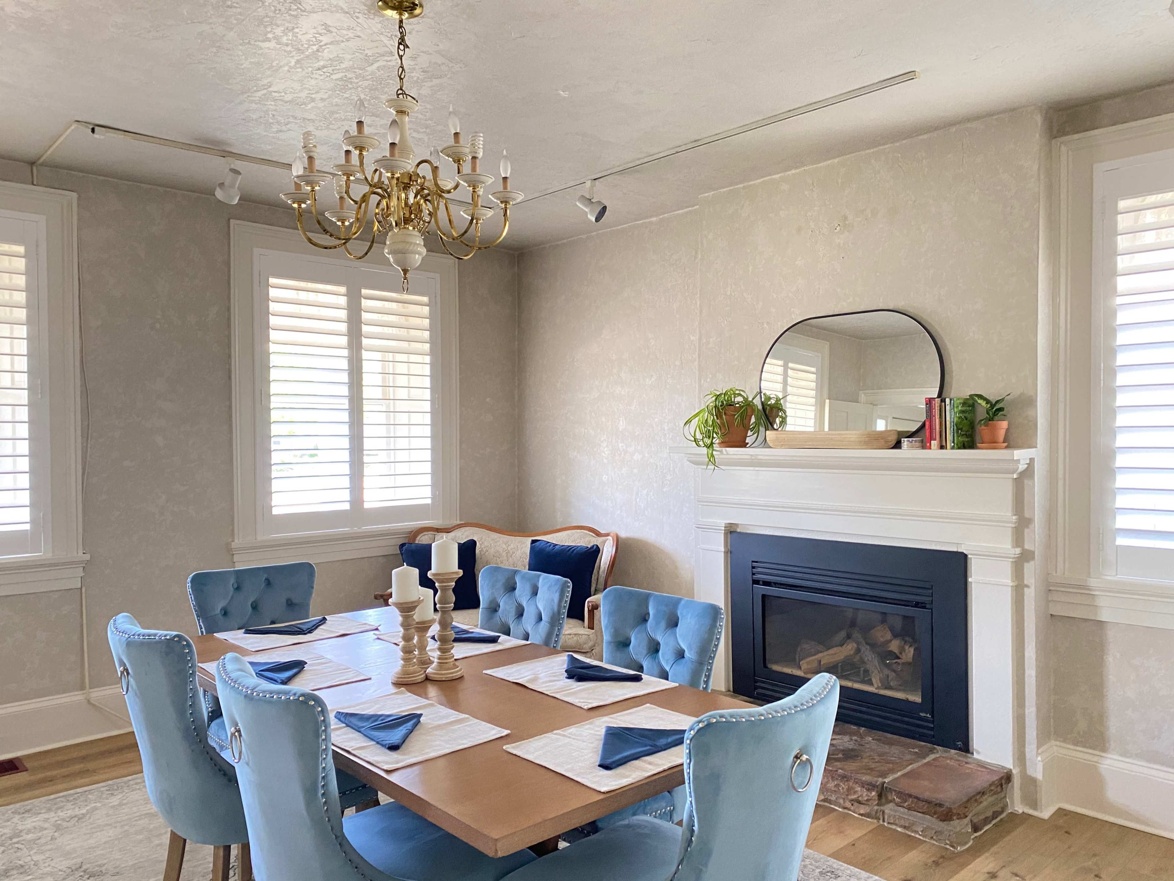 A dining room features a table set for six under a chandelier, with a fireplace and large windows adorned with plantation shutters.