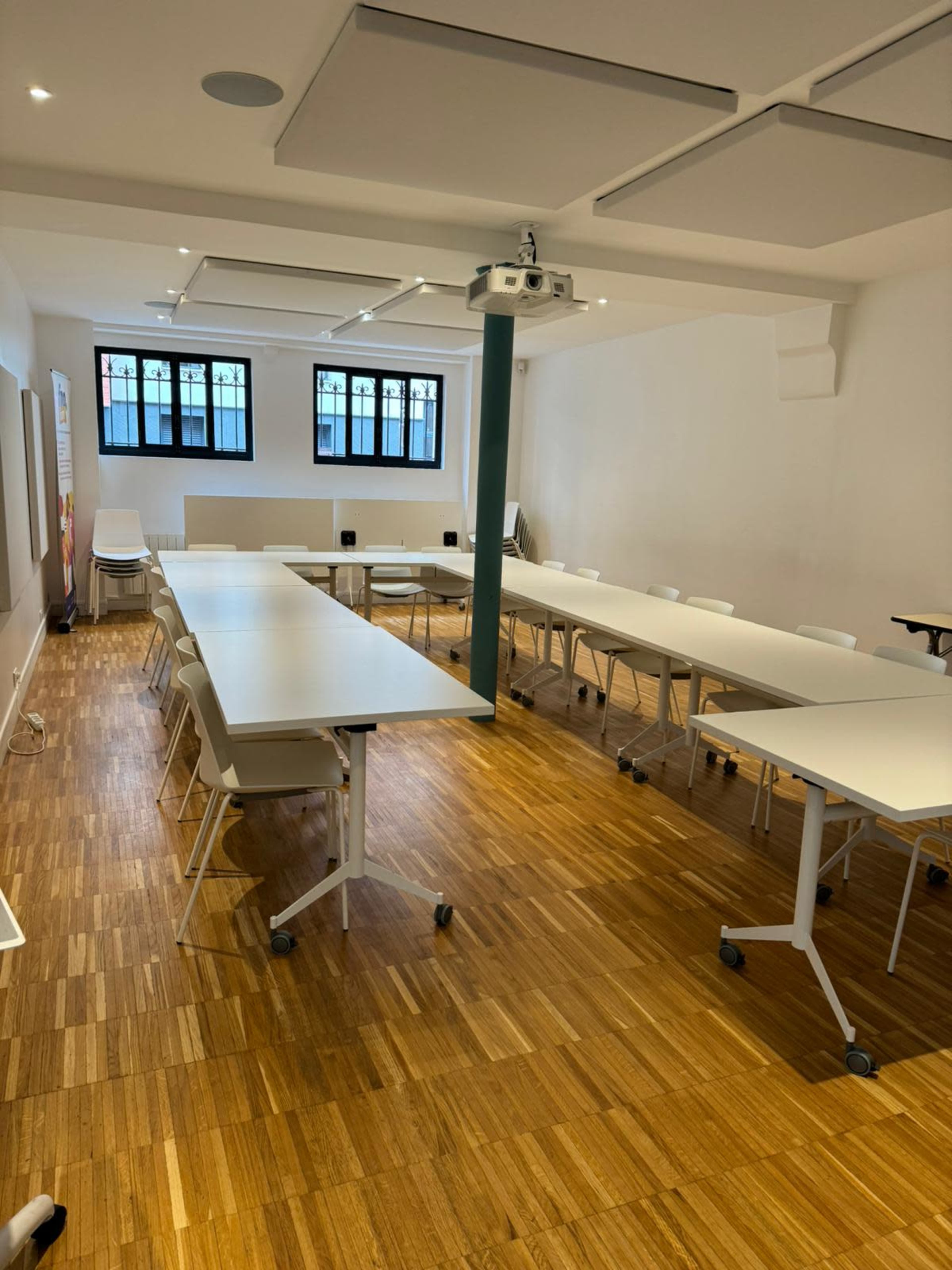 A conference room features multiple rectangular tables arranged in a U-shape, with white chairs and a projector on the ceiling.