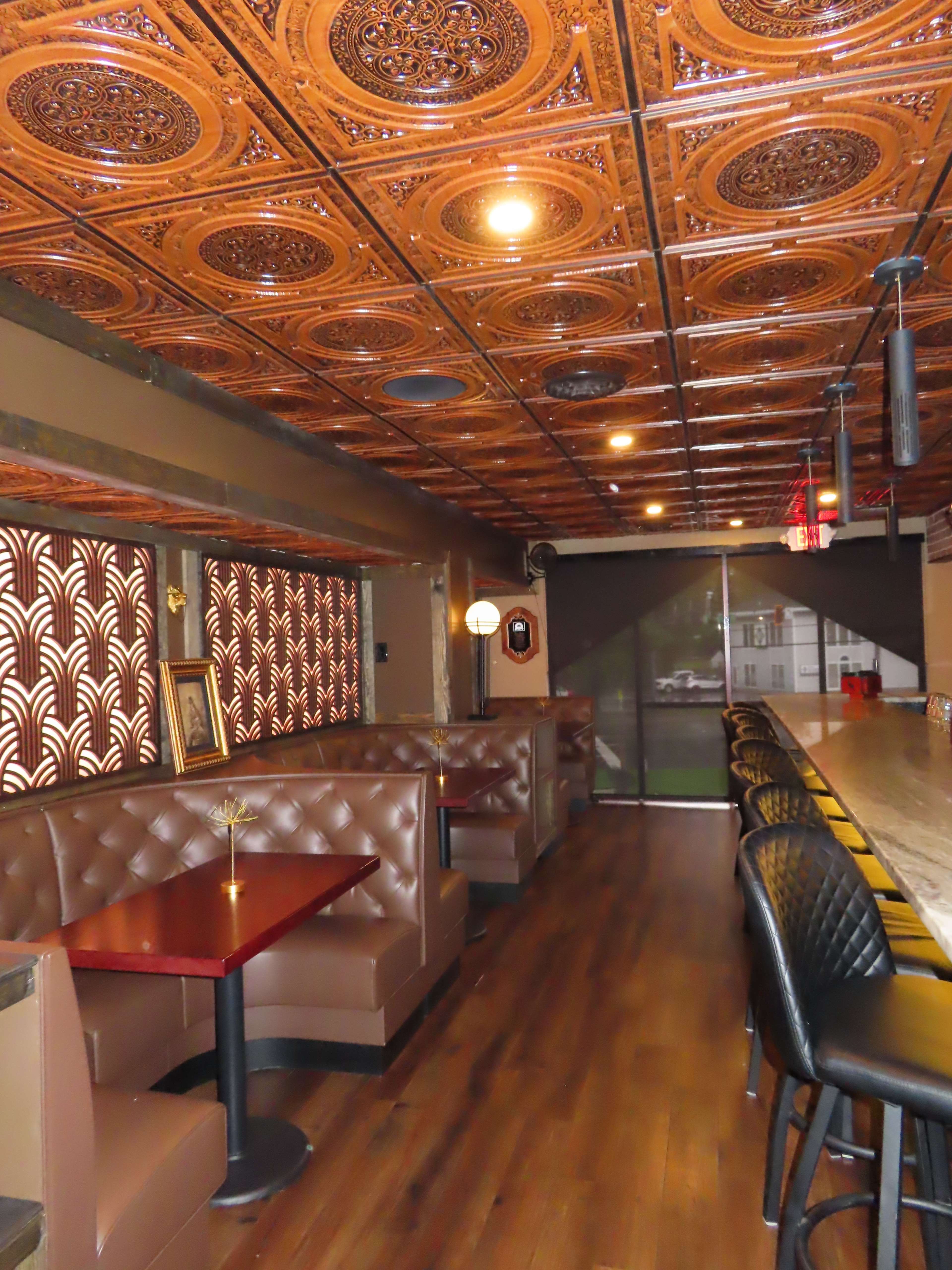 The image shows an interior view of a restaurant featuring leather booth seating, a long bar with black chairs, and an ornate wooden ceiling.