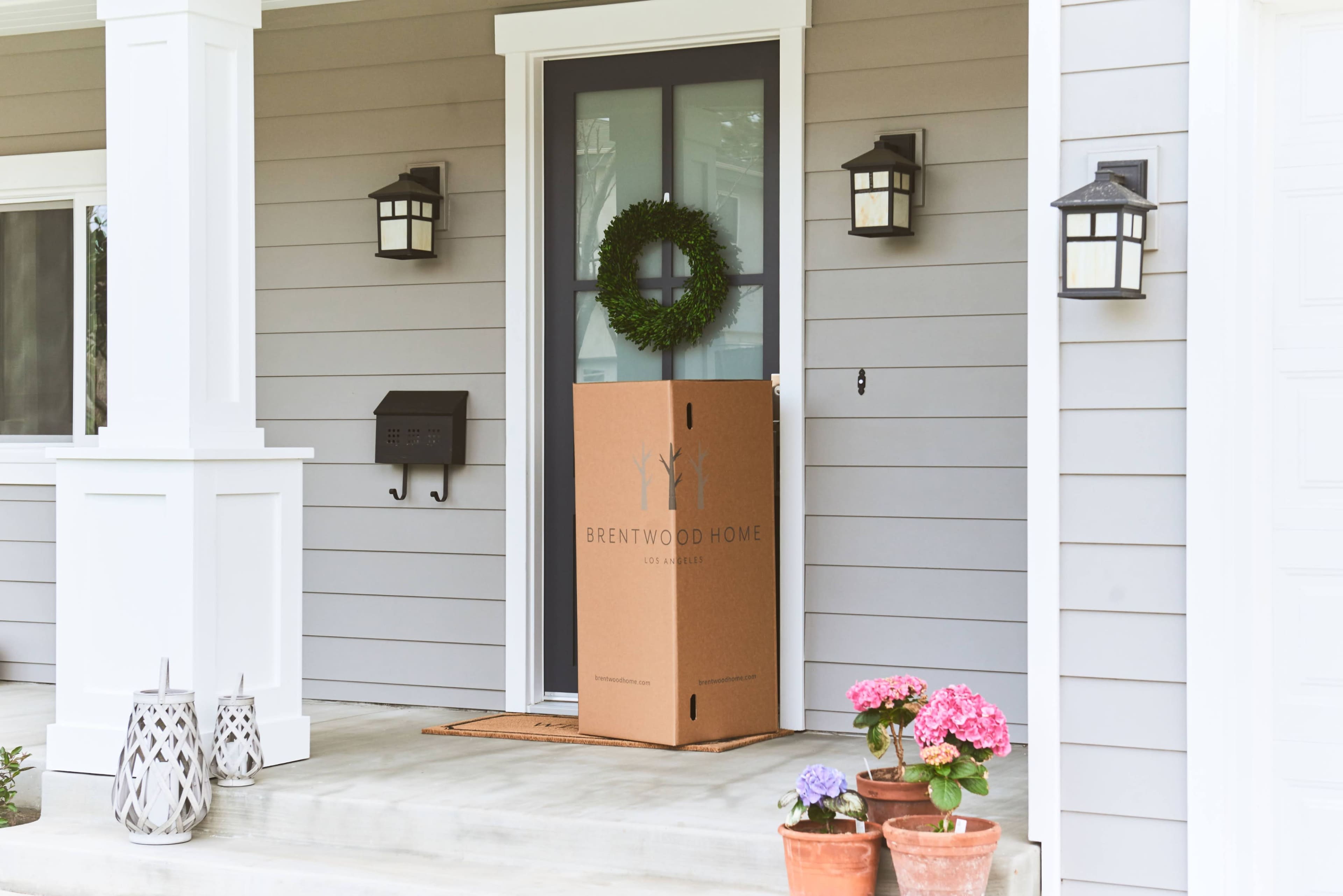 A large cardboard box with the logo "Brentwood Home" sits on a porch beside potted flowers and a wreath on the door.
