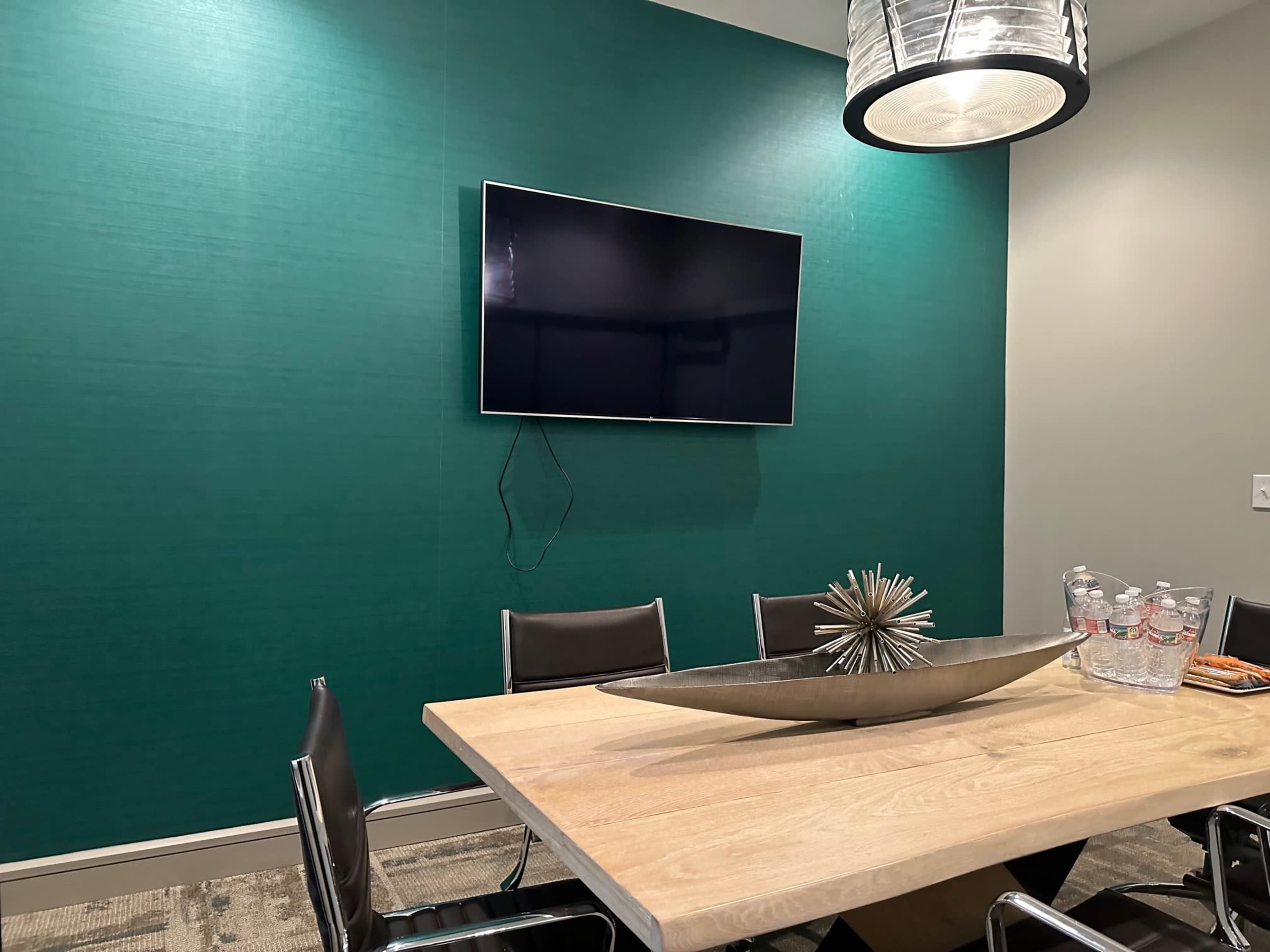 The image shows a conference room with a wooden table, black chairs, a large decorative centerpiece, a TV mounted on a teal wall, and a set of water bottles placed off to the side.
