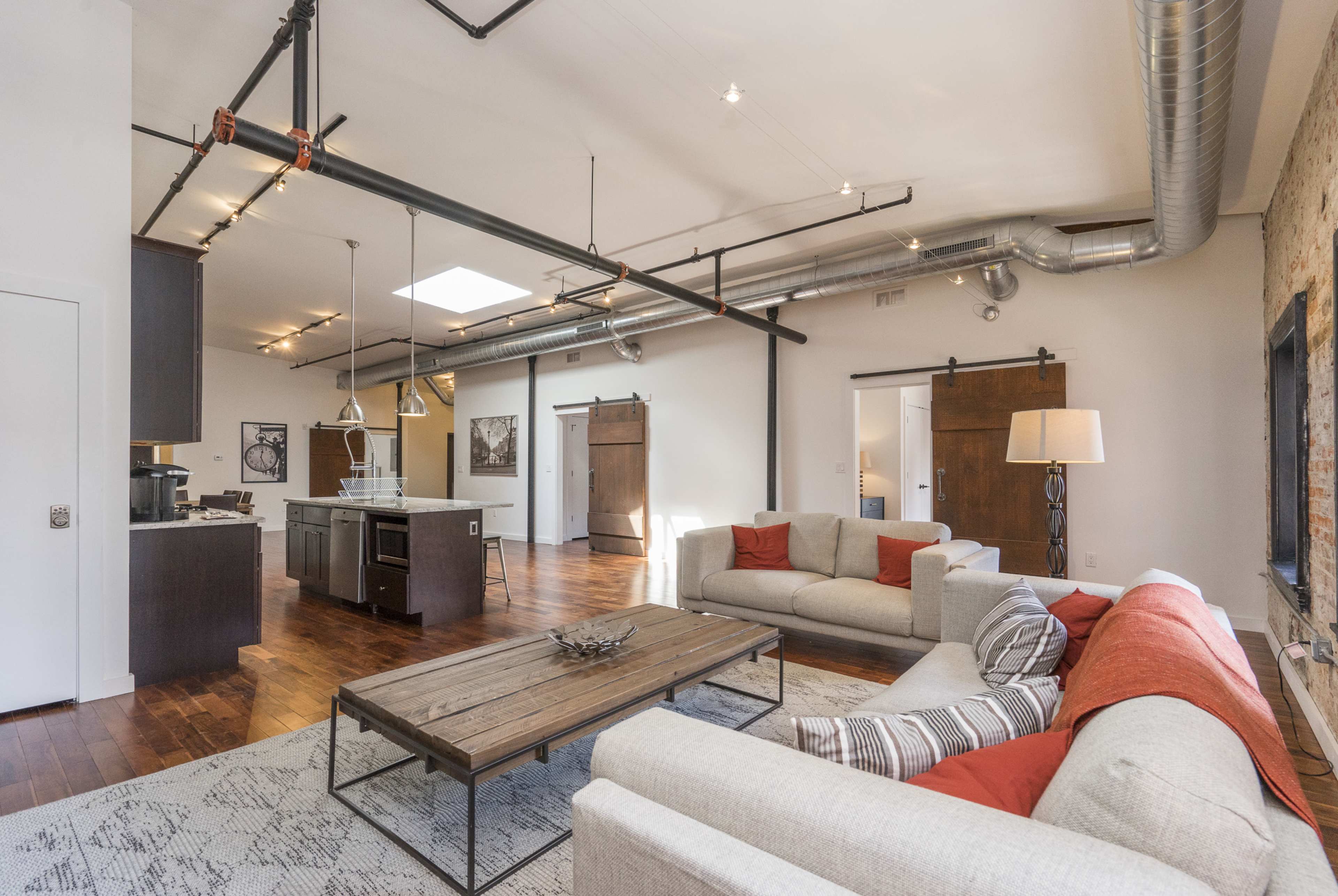 A spacious, modern living area with a kitchen to the left, featuring hardwood floors, industrial piping, and large sliding barn doors.