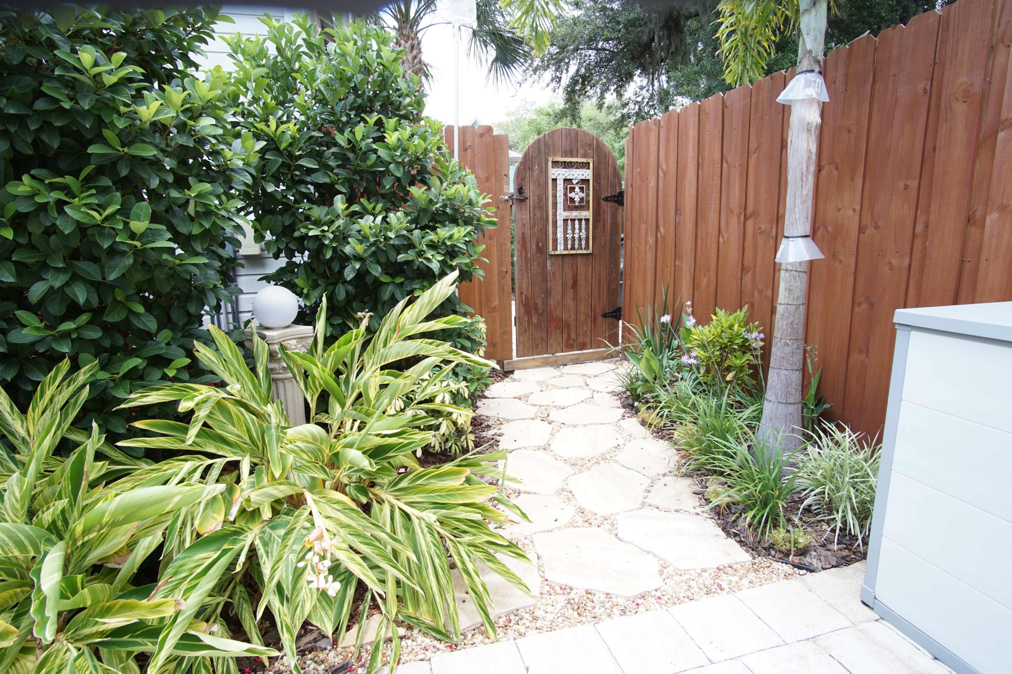 A stone pathway leads to a wooden gate surrounded by lush greenery and a fenced area.