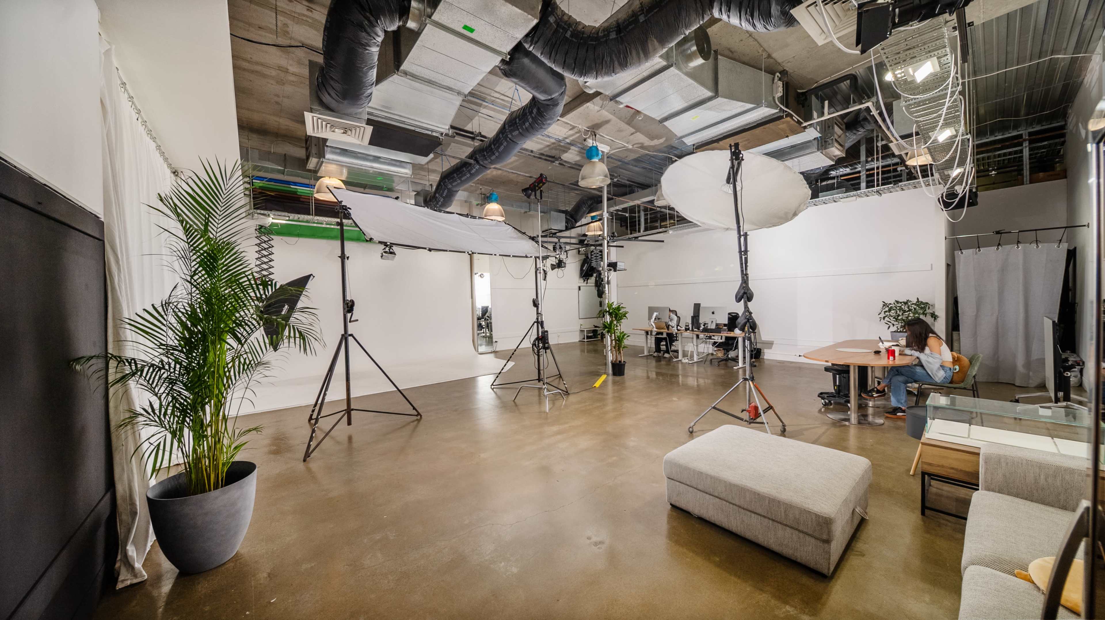 The image shows a spacious studio setting with professional lighting equipment, a sofa, a desk area, and a person seated at a table in front of a white backdrop.