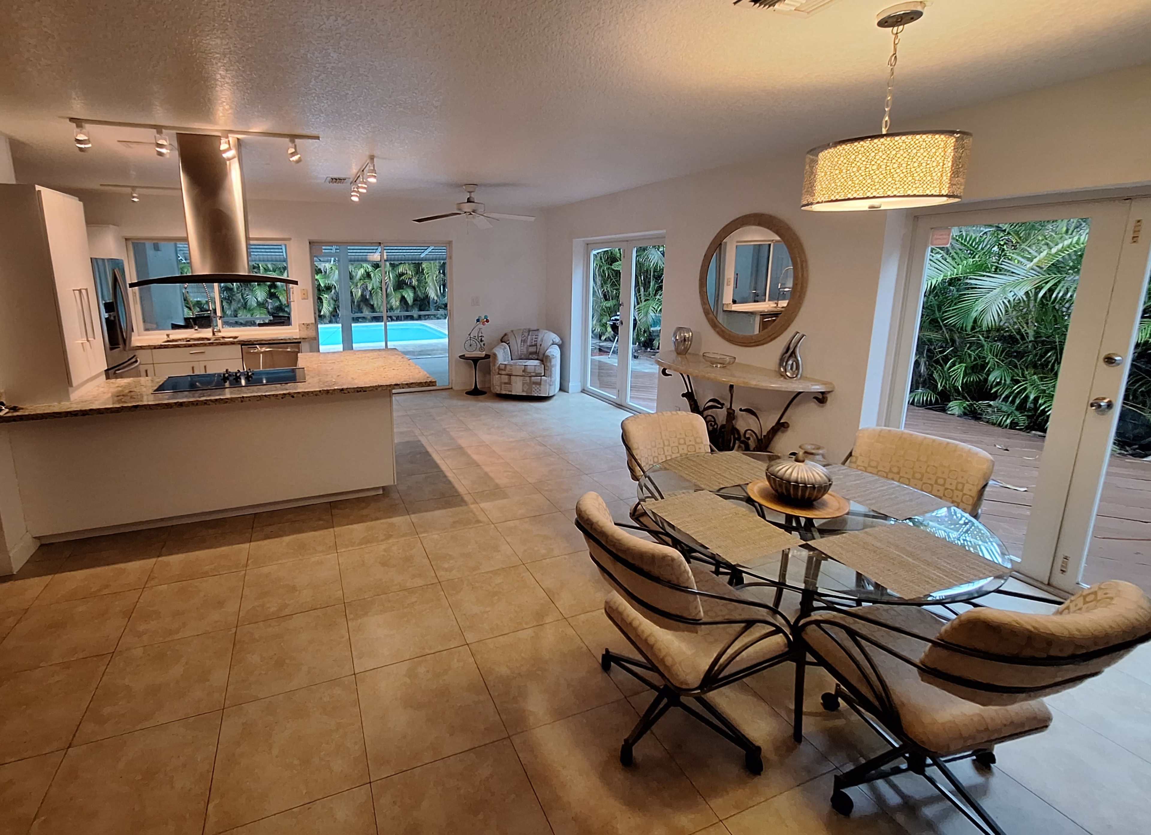 An open-concept kitchen and dining area with a tiled floor, a round dining table, and large windows that overlook a pool area surrounded by greenery.
