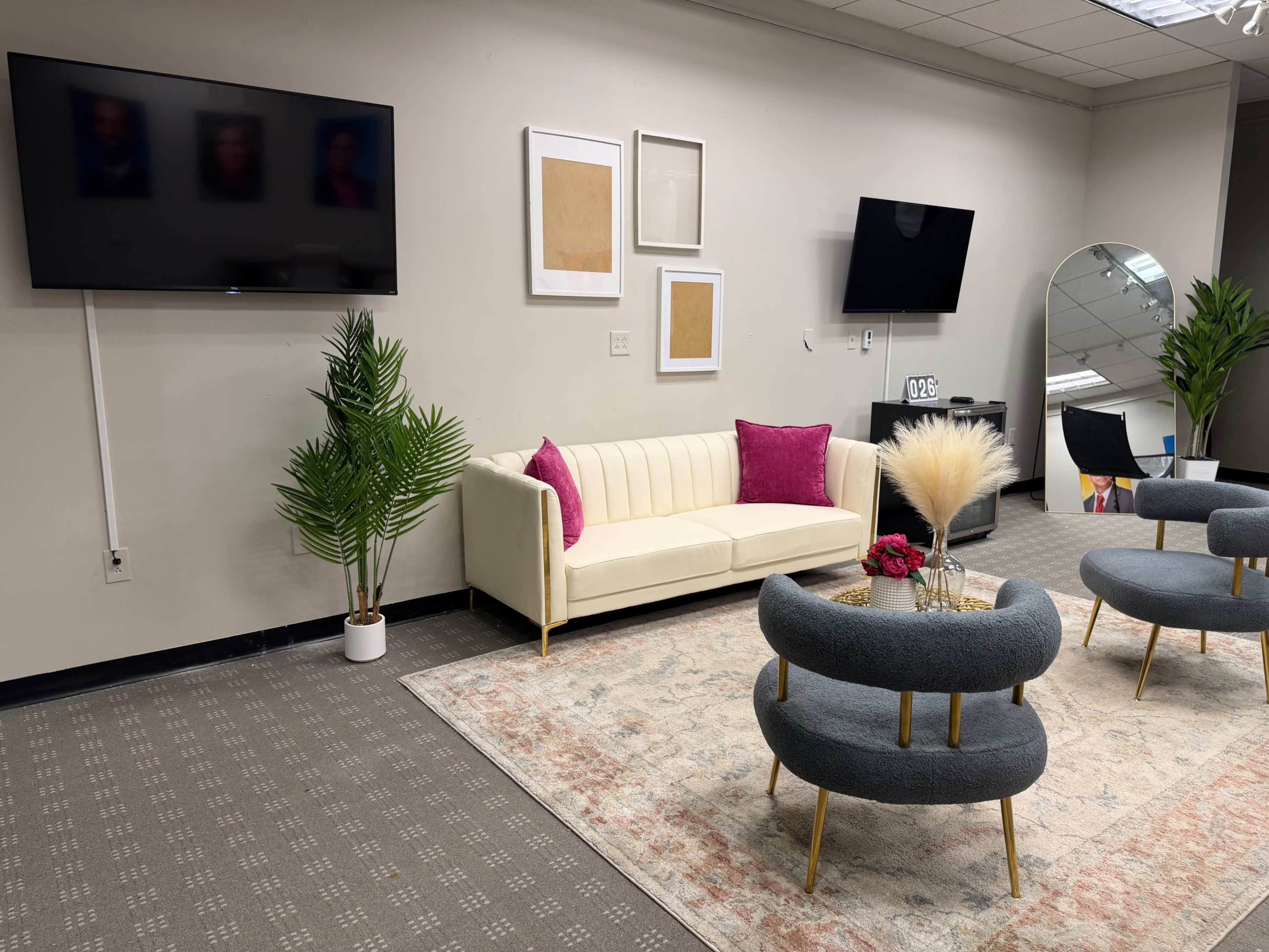 The image shows a modern, well-lit lounge area featuring a light-colored couch with pink pillows, two curved gray chairs, a round table with decorative elements, and two wall-mounted flatscreen TVs.