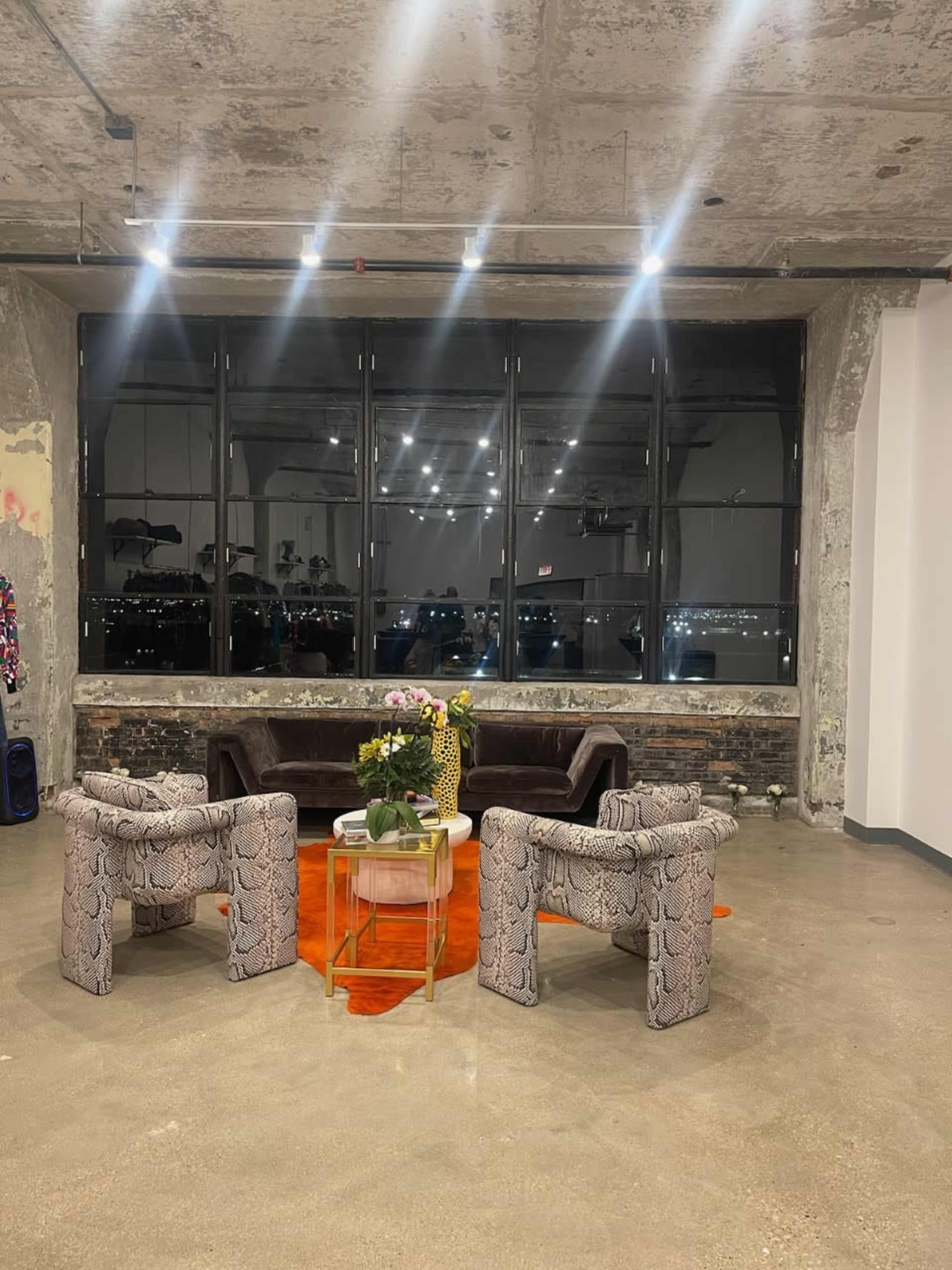 The image shows a modern living area with a dark sofa, two patterned armchairs, a small table with flowers, and large windows displaying a city view at night.