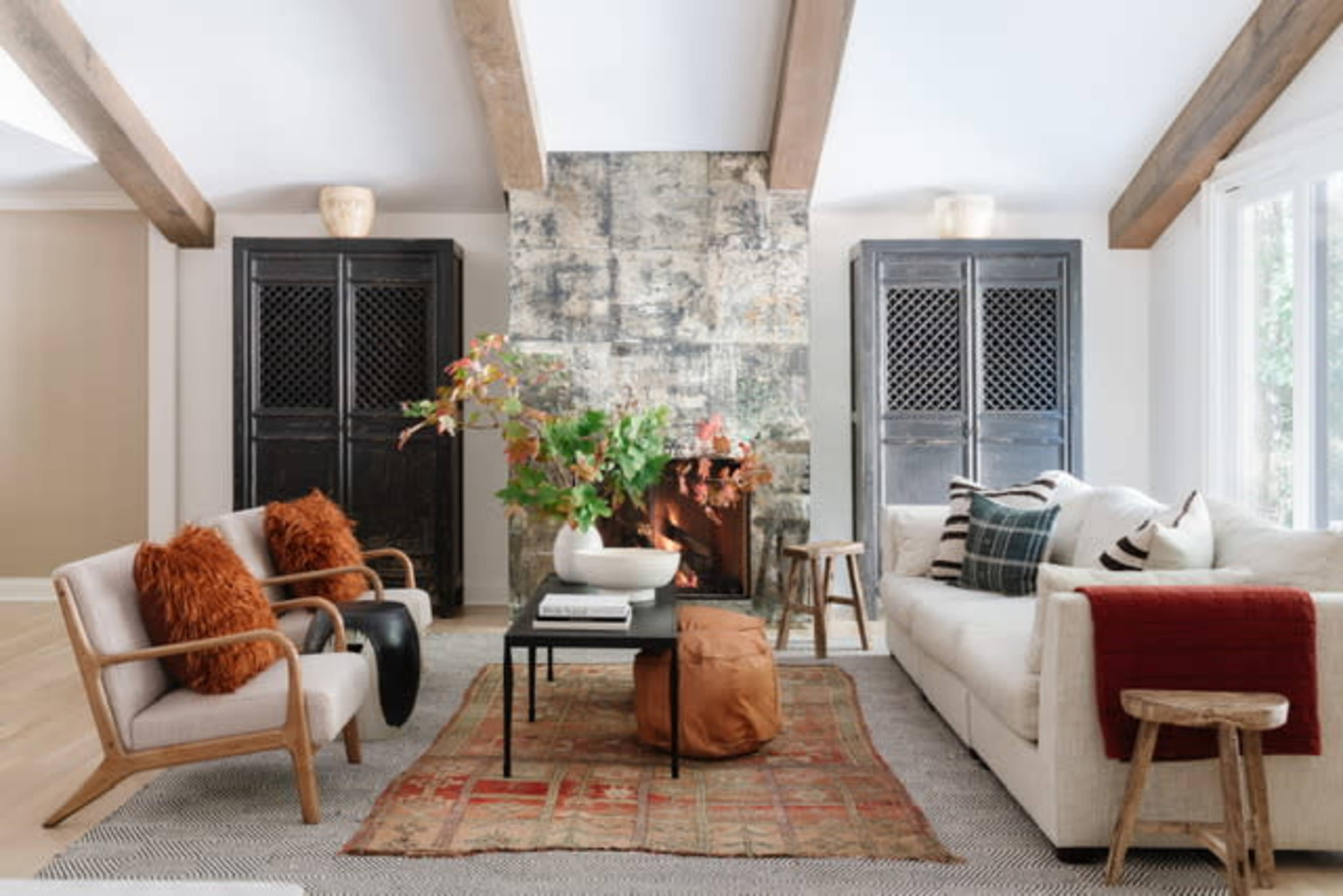 The living room features a stone fireplace with a floral arrangement on a coffee table, flanked by two dark cabinets, cozy seating, and a patterned area rug.