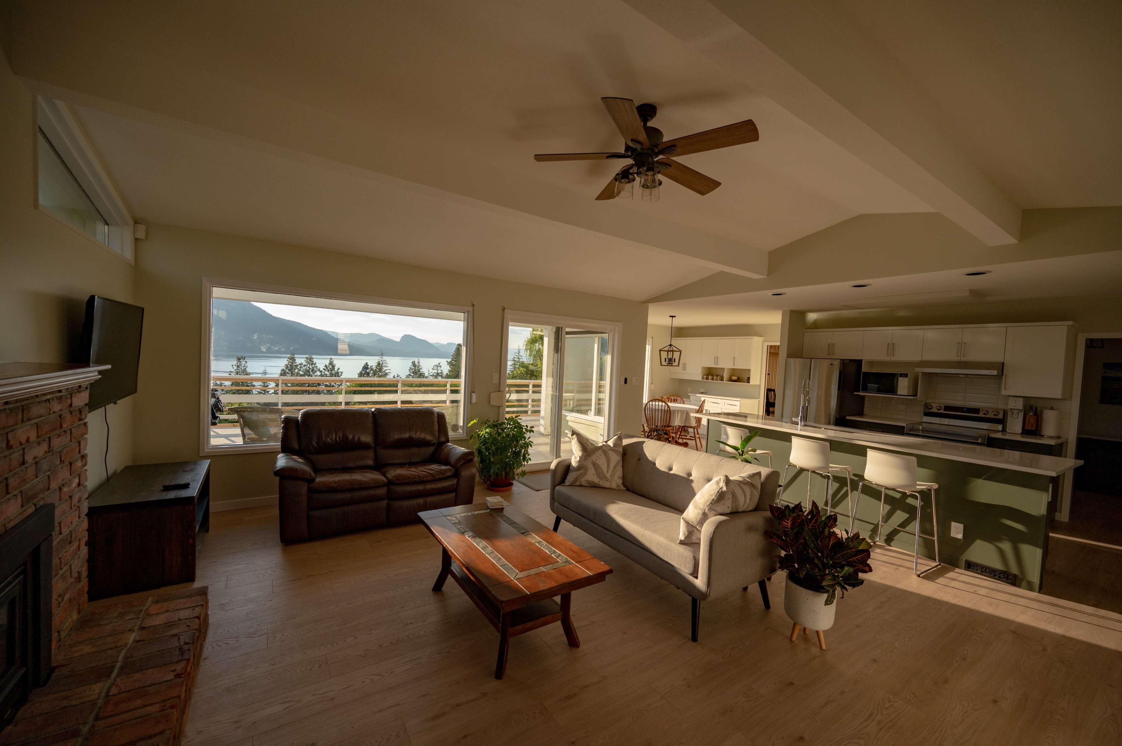 The interior of a modern living room features a sofa and armchair facing a coffee table, with large windows providing a view of a lake and mountains.