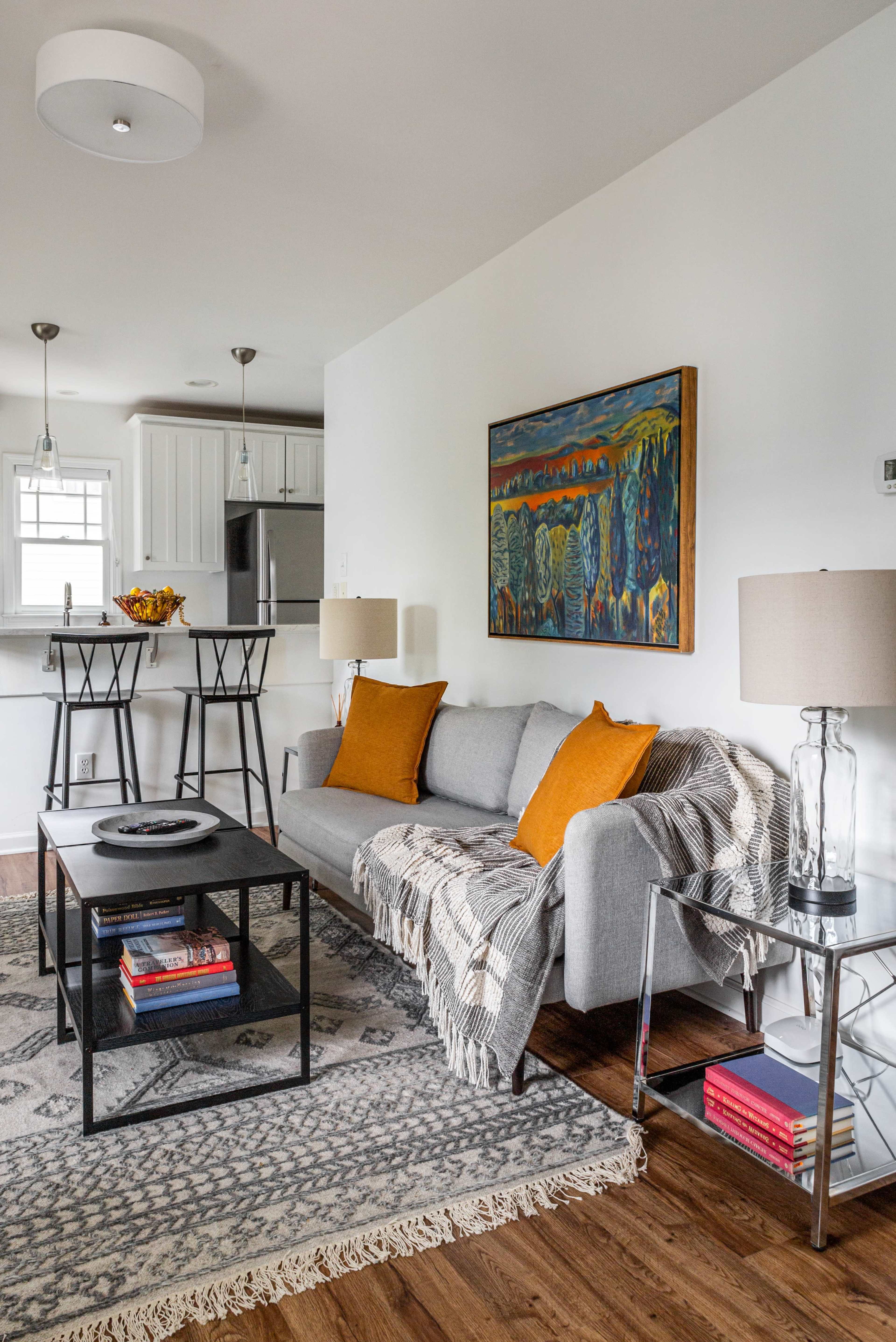 A modern living room with a gray sofa, orange cushions, a black coffee table, and a large painting on the wall, complemented by a patterned rug and two lamps on side tables.