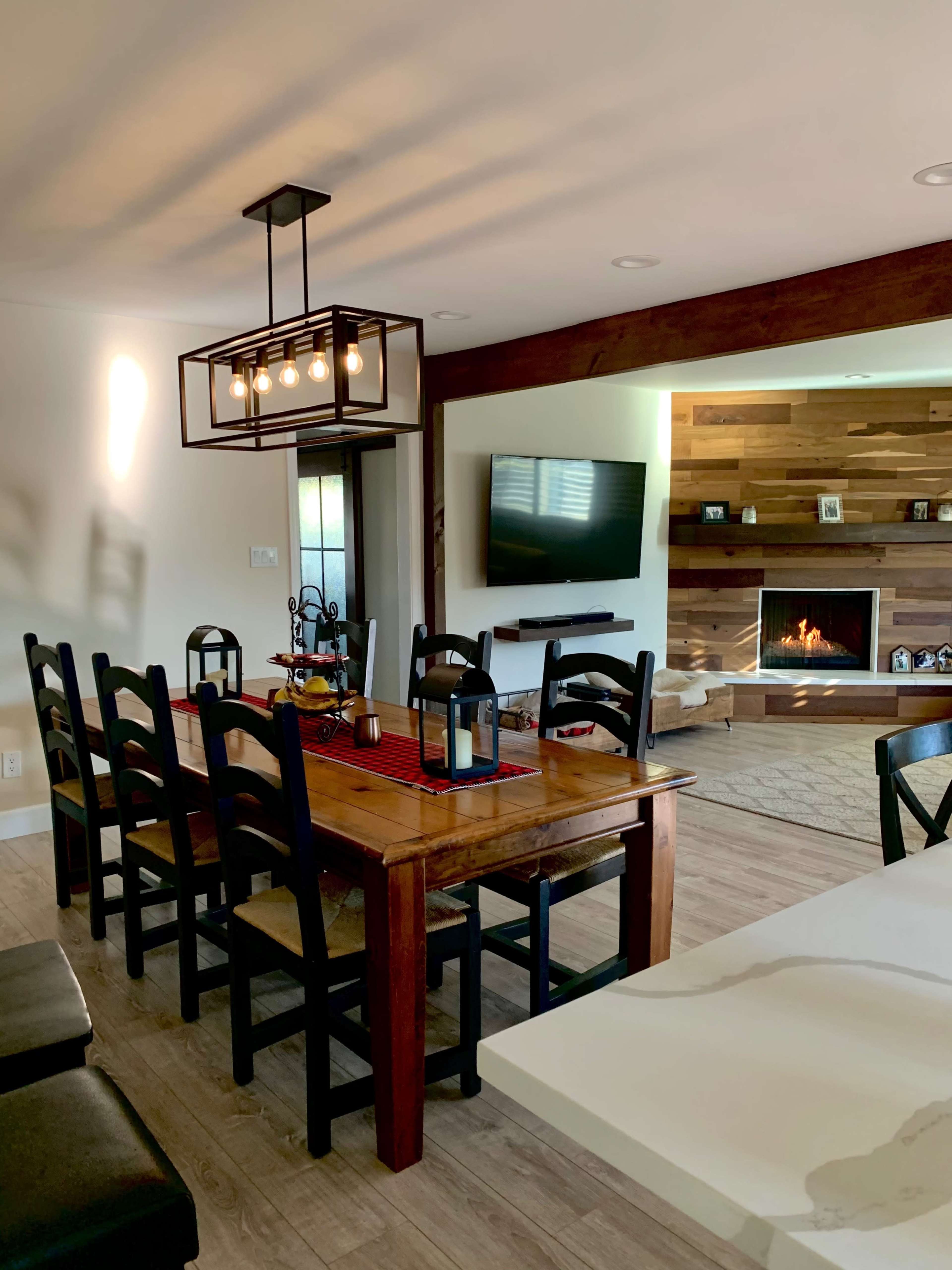 A dining area features a wooden table with black chairs, a modern light fixture above, and an adjacent living space with a fireplace and TV.