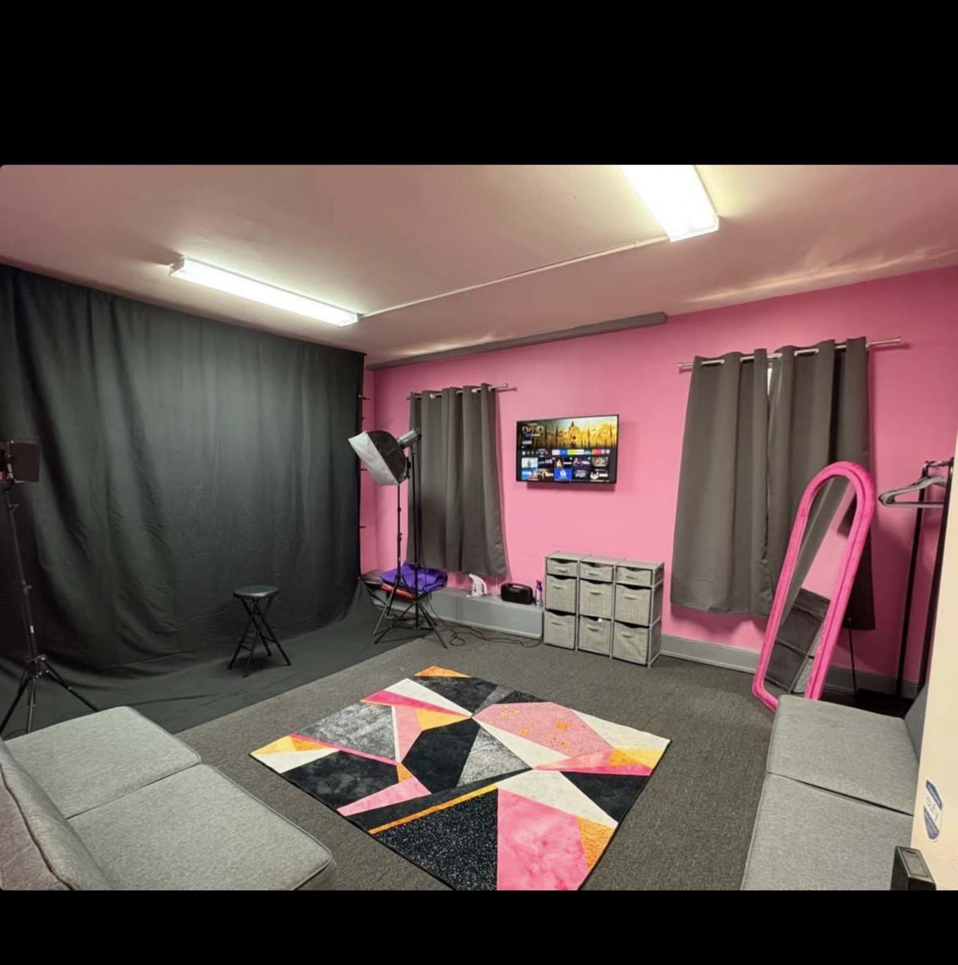 The image shows a small studio space with pink walls, a large black curtain, a television mounted on the wall, a gray rug with geometric patterns, and seating areas.