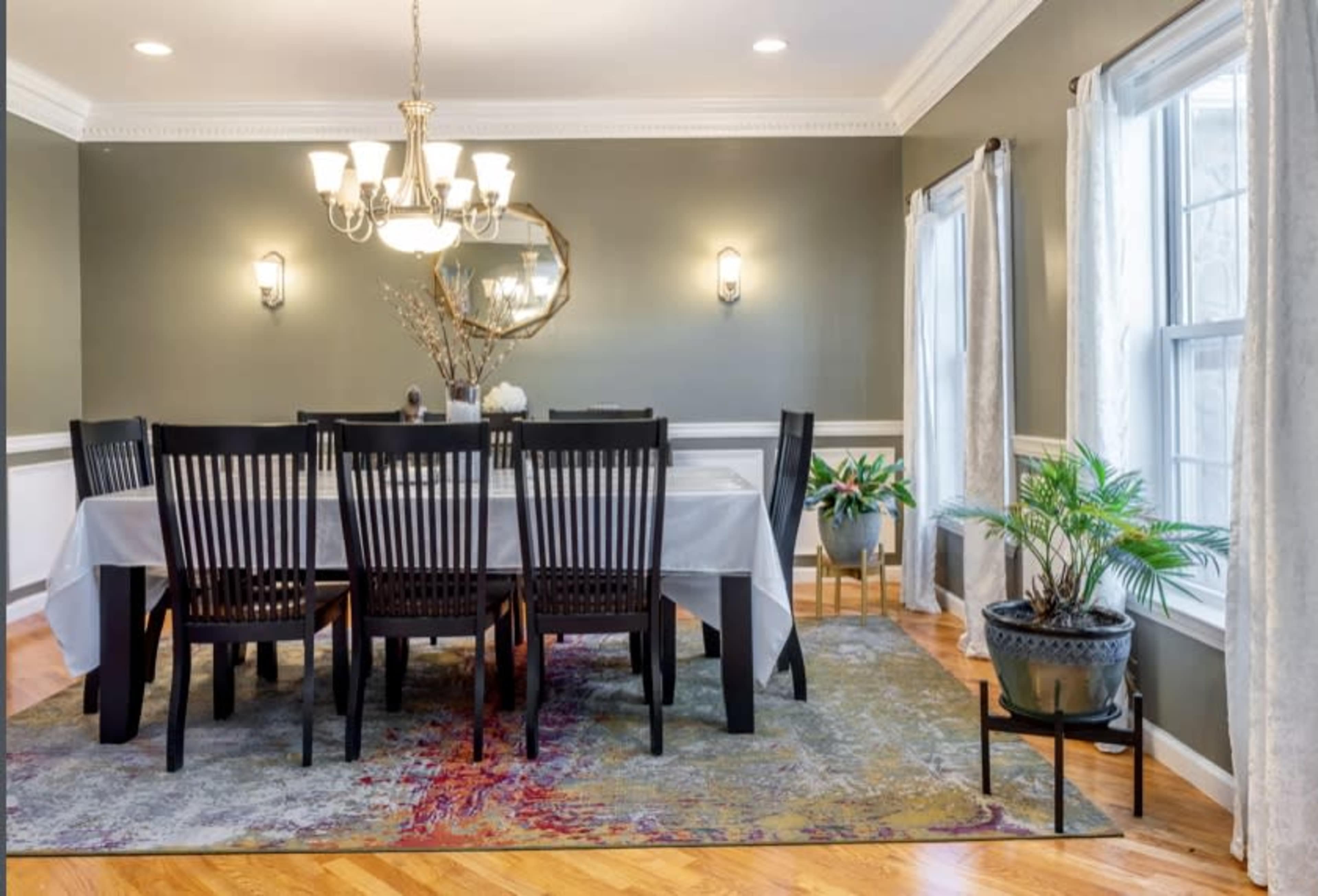 A dining room features a long table surrounded by black chairs, with a chandelier above and large windows allowing natural light to enter.