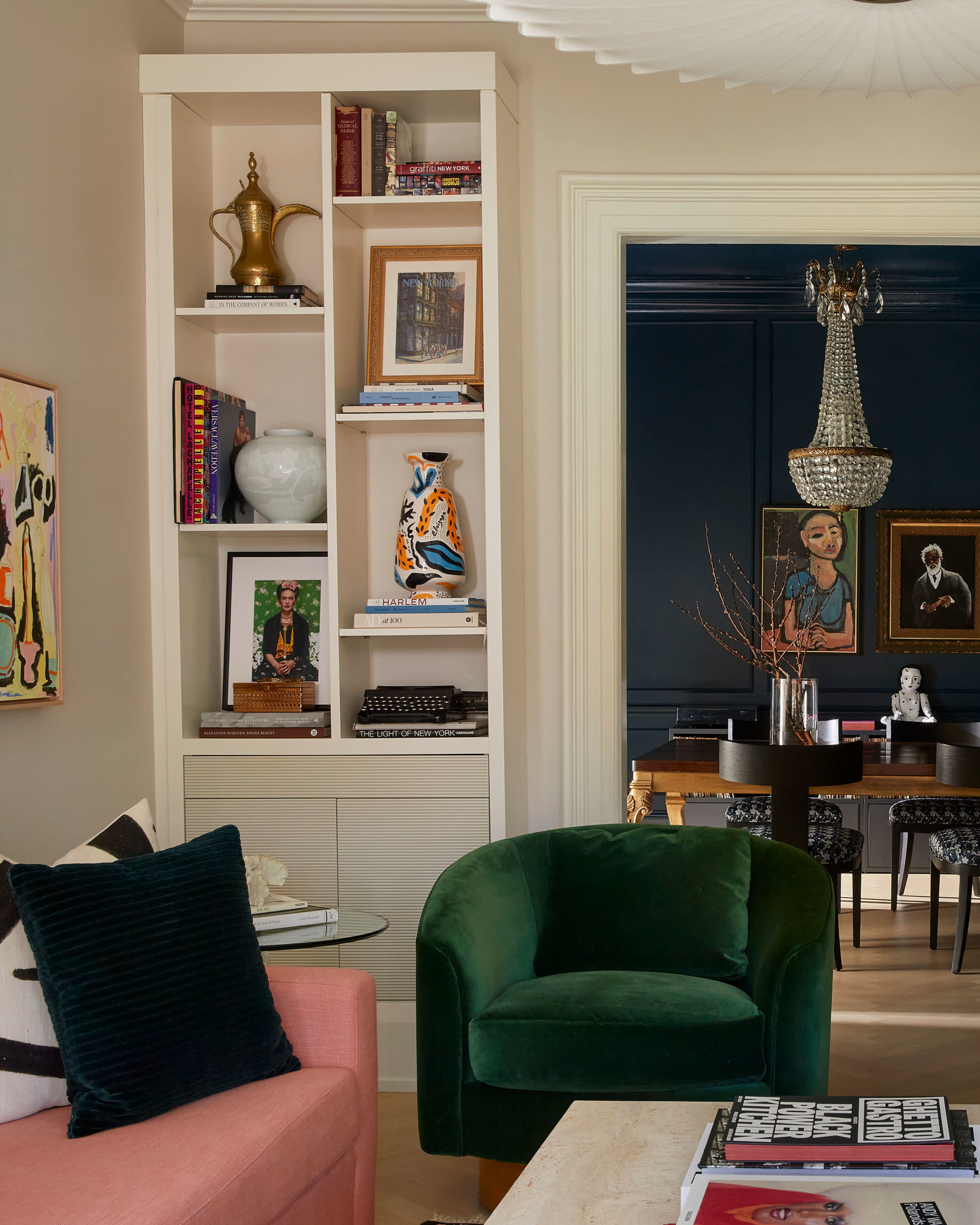 The image shows a living room with a pink sofa, a green armchair, and a built-in shelving unit displaying various books and decor items.