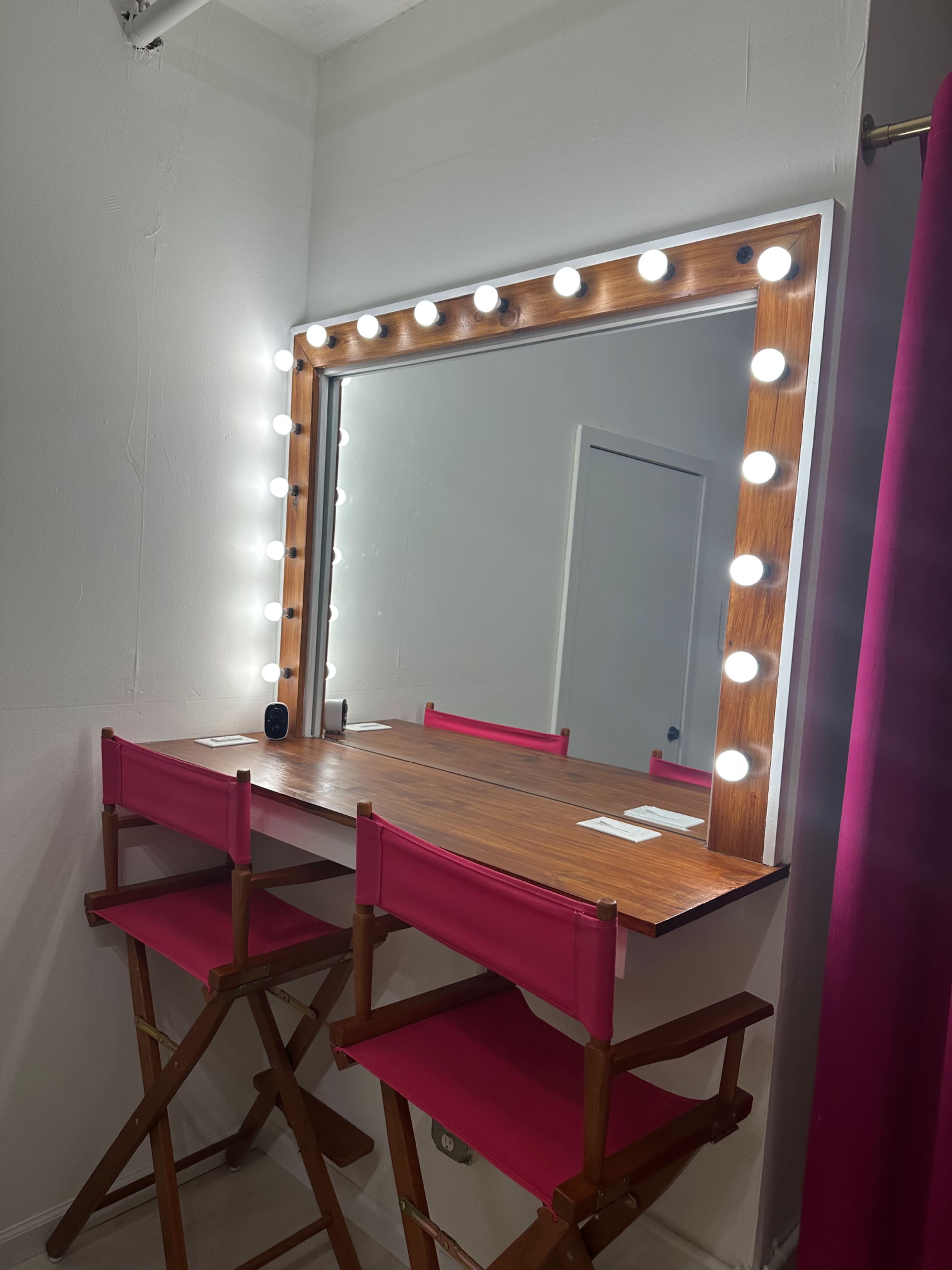 A well-lit vanity mirror with Hollywood-style lights is positioned above a wooden table, accompanied by two pink director's chairs.