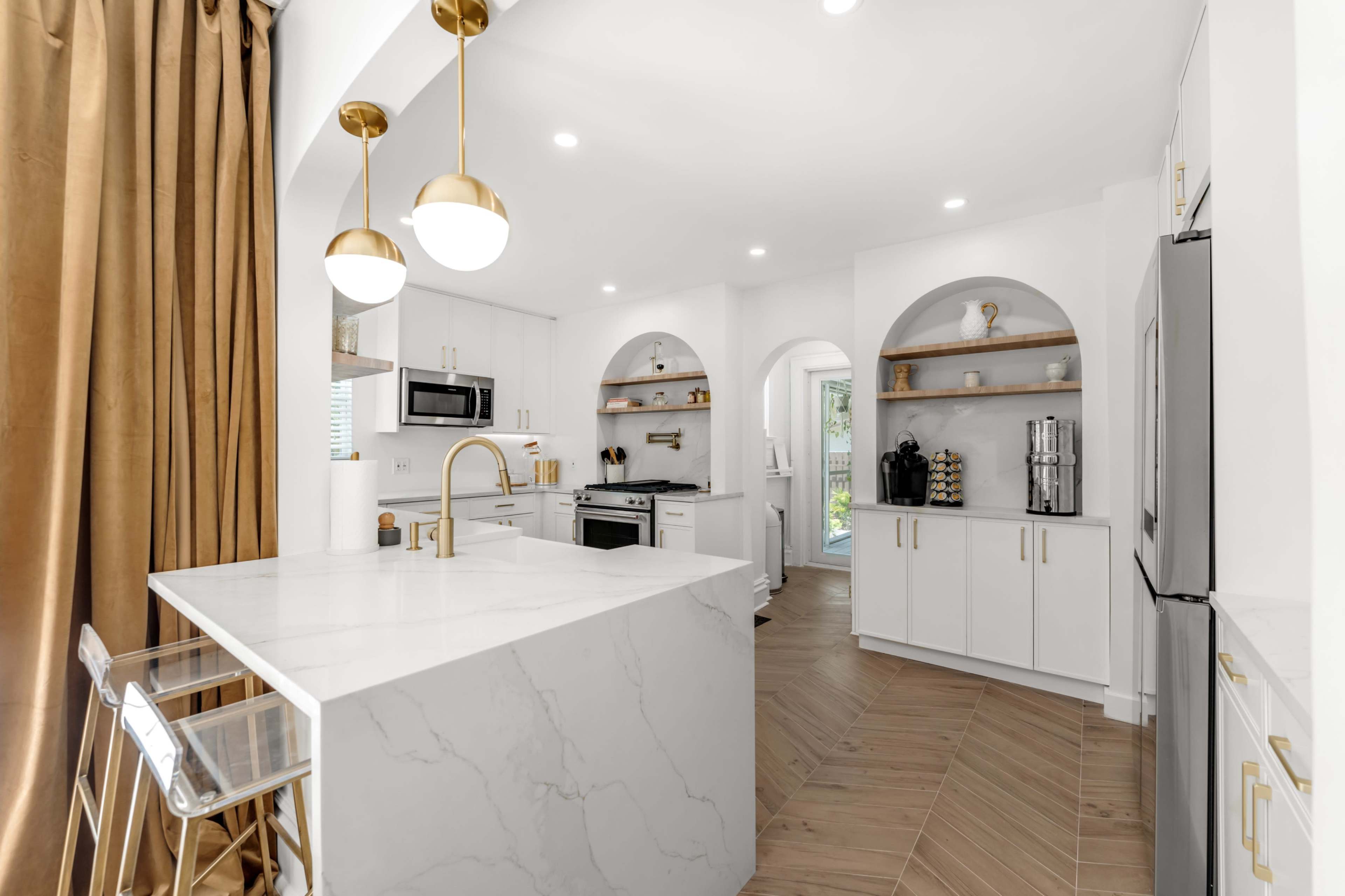 The image shows a modern kitchen featuring white cabinetry, a large marble island, gold pendant lights, and wooden flooring.