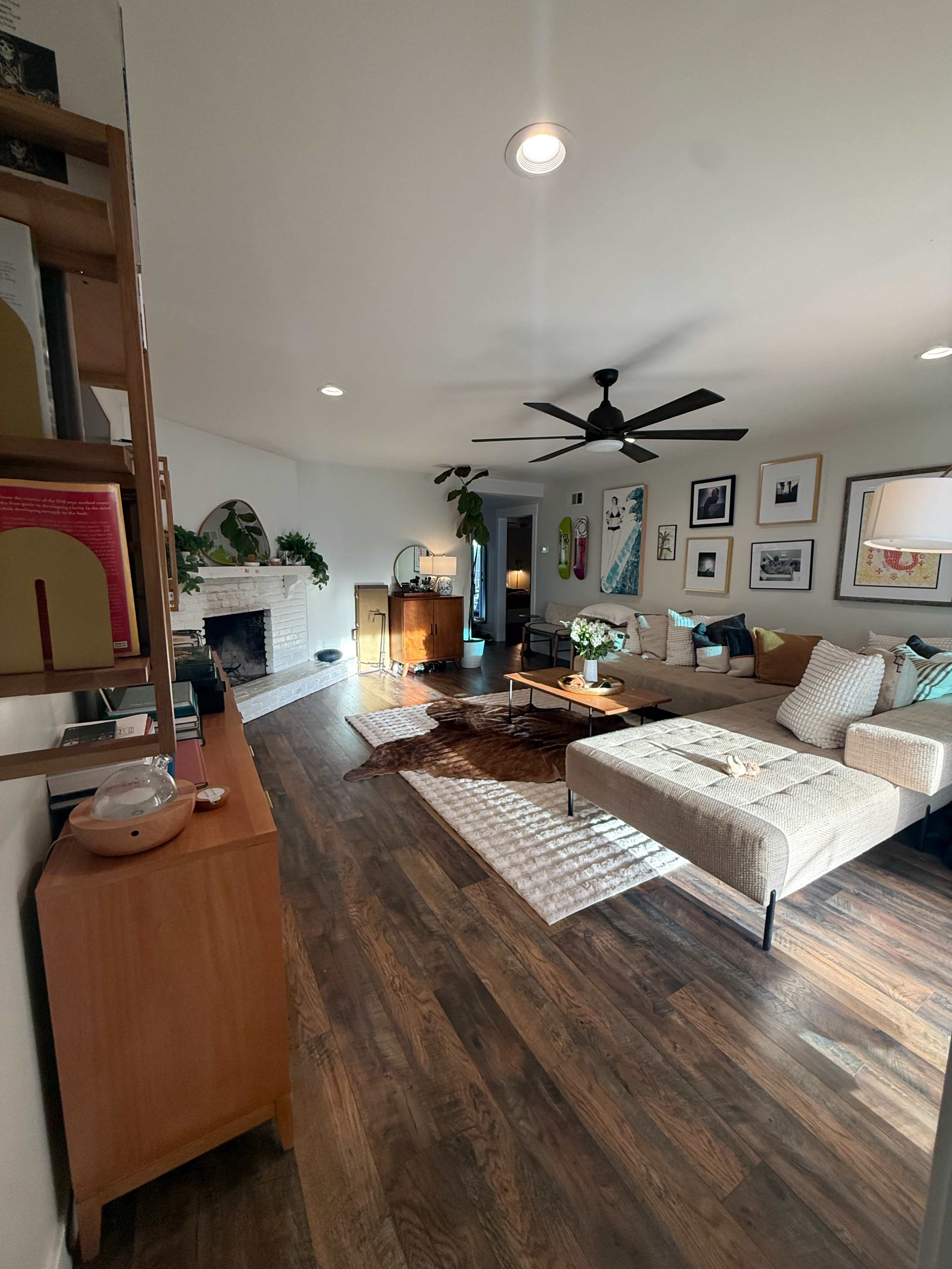 The image shows a living room with a beige sectional sofa, a coffee table, a fireplace, and various framed pictures on the walls, all set against a backdrop of wooden flooring.