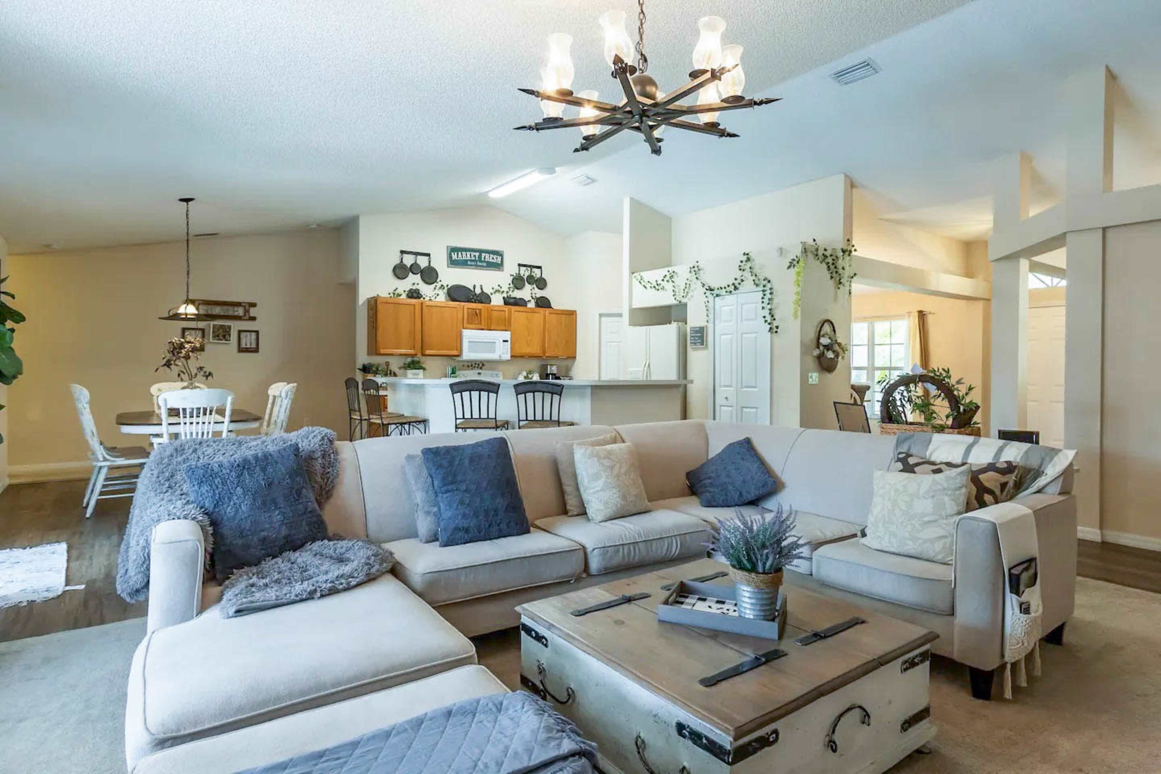 A spacious living room features a light-colored sectional sofa, a wooden coffee table, and an open layout connecting to a dining area and kitchen.