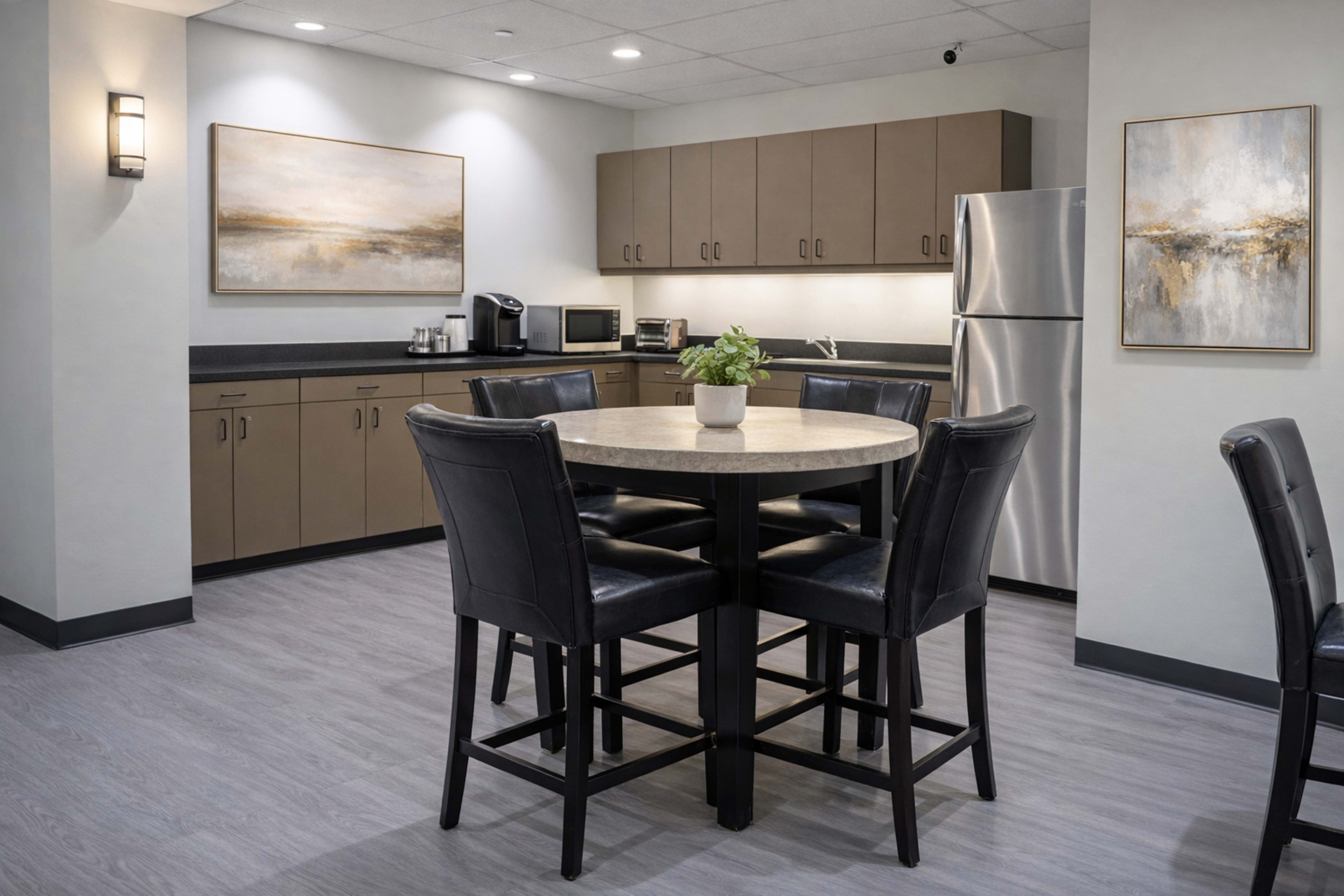 The image shows a modern kitchen area with a circular dining table surrounded by tall chairs, beige cabinets, and stainless steel appliances.