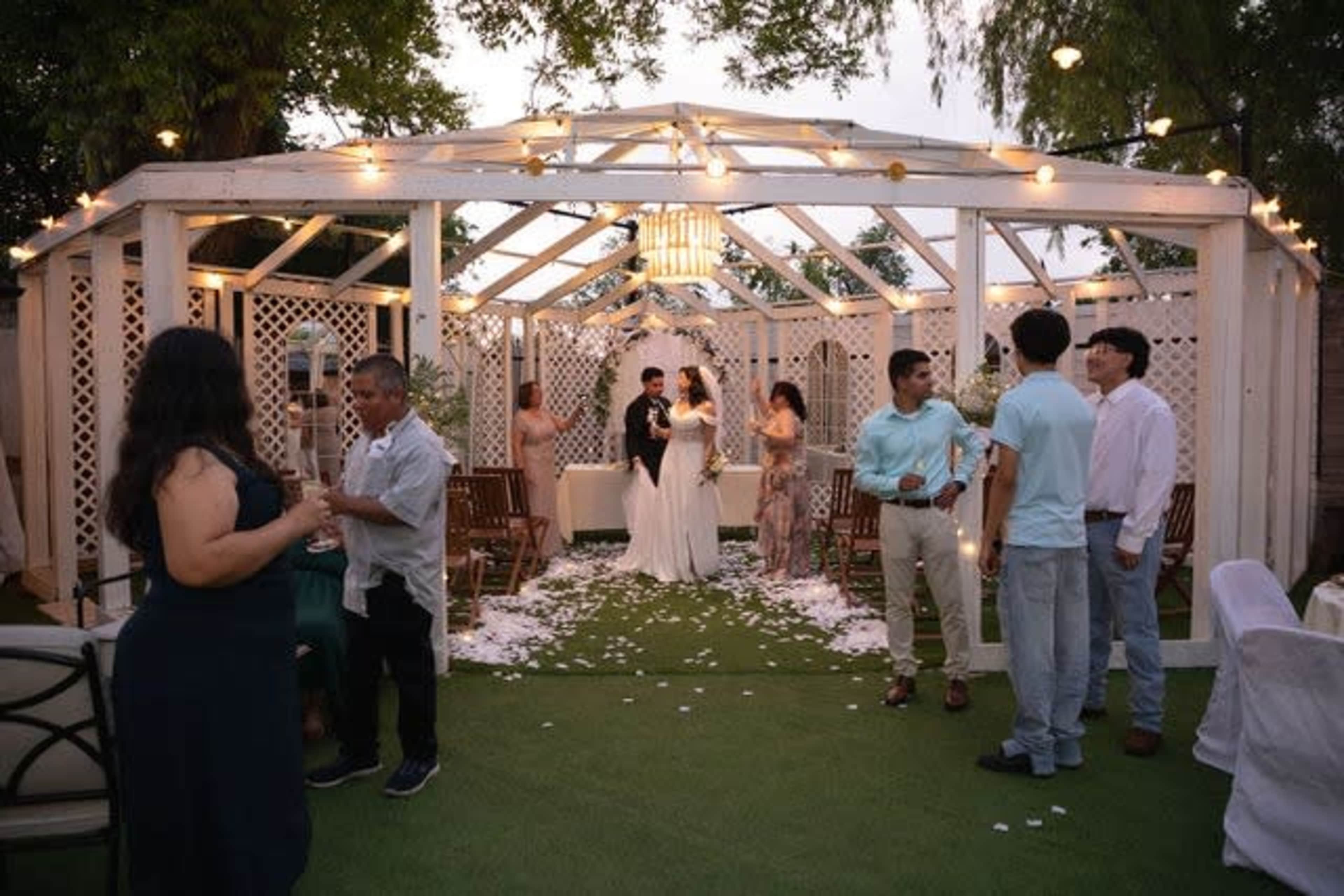 A wedding ceremony is taking place under a decorated white gazebo, surrounded by guests and adorned with hanging lights and scattered flower petals.