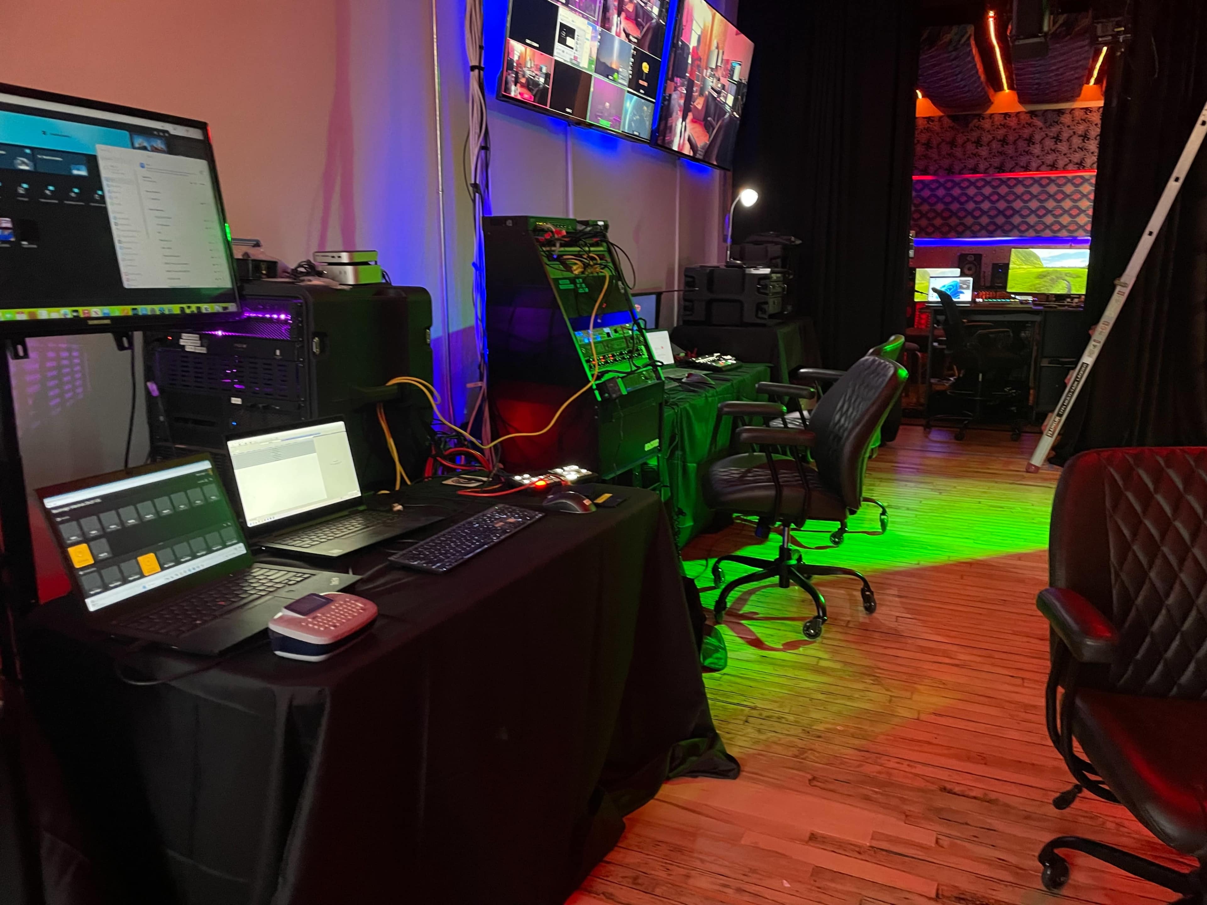 The image shows a view of a control room with multiple screens displaying digital content, along with computers, cables, and chairs arranged in a workspace.